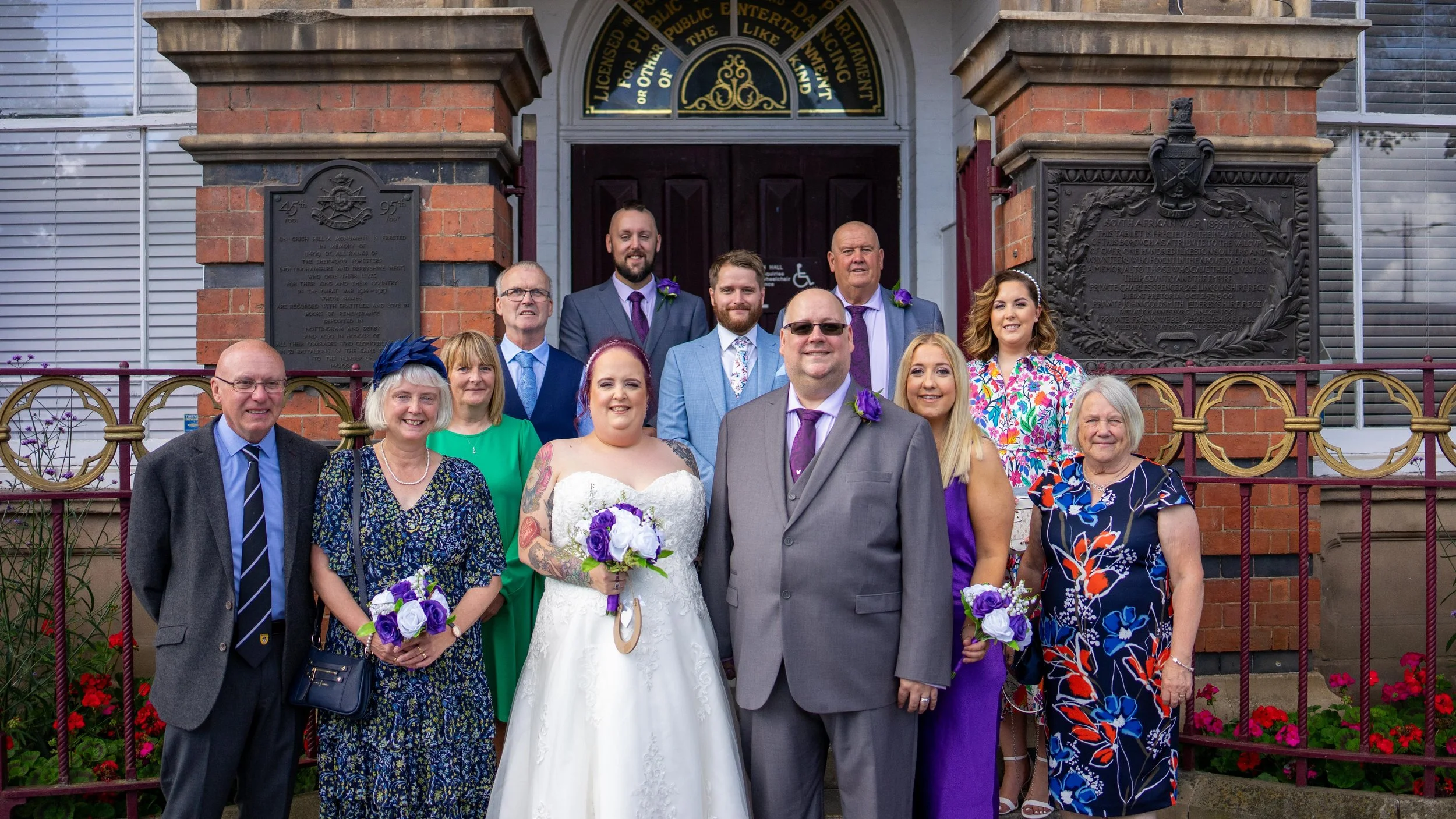 A wedding group photo outside a brick church showing a bride in a white dress holding a bouquet, surrounded by family and friends in colorful attire.