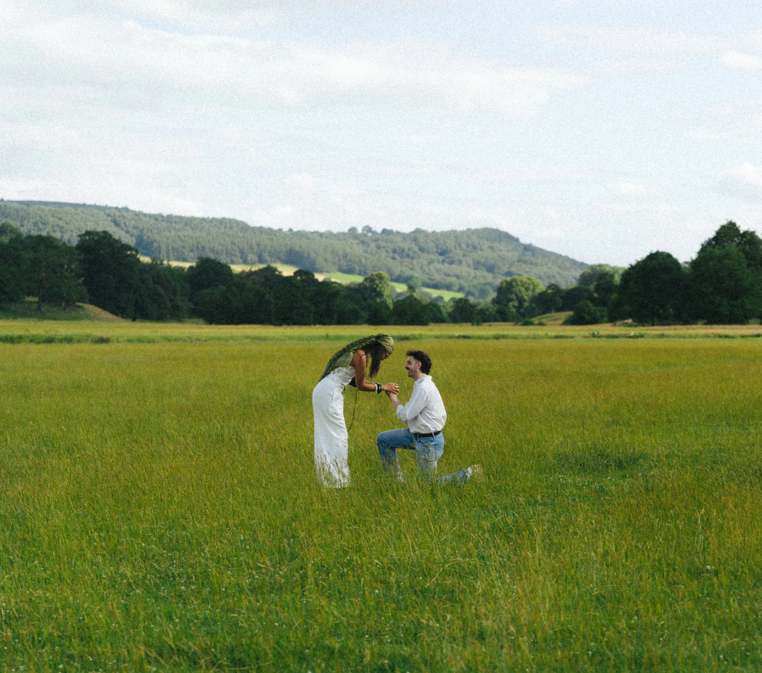 A couple kneeling in a green field, holding hands and smiling at each other with hills and trees in the background.