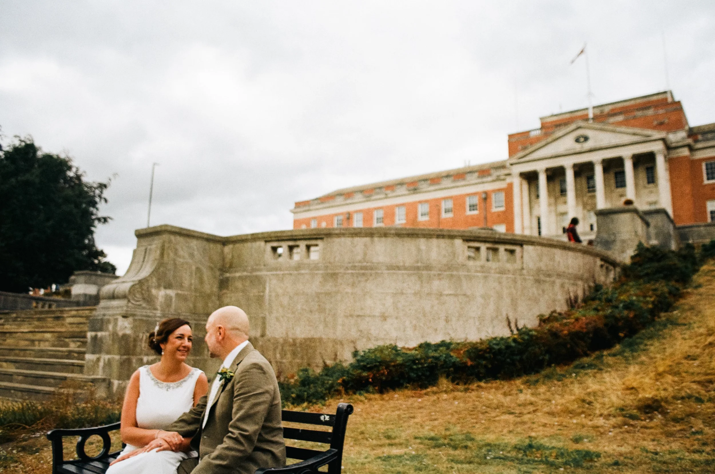 A couple sitting on a park bench at Chesterfield after their wedding, smiling at each other during an outdoor wedding, with a historic building and stairs in the background.