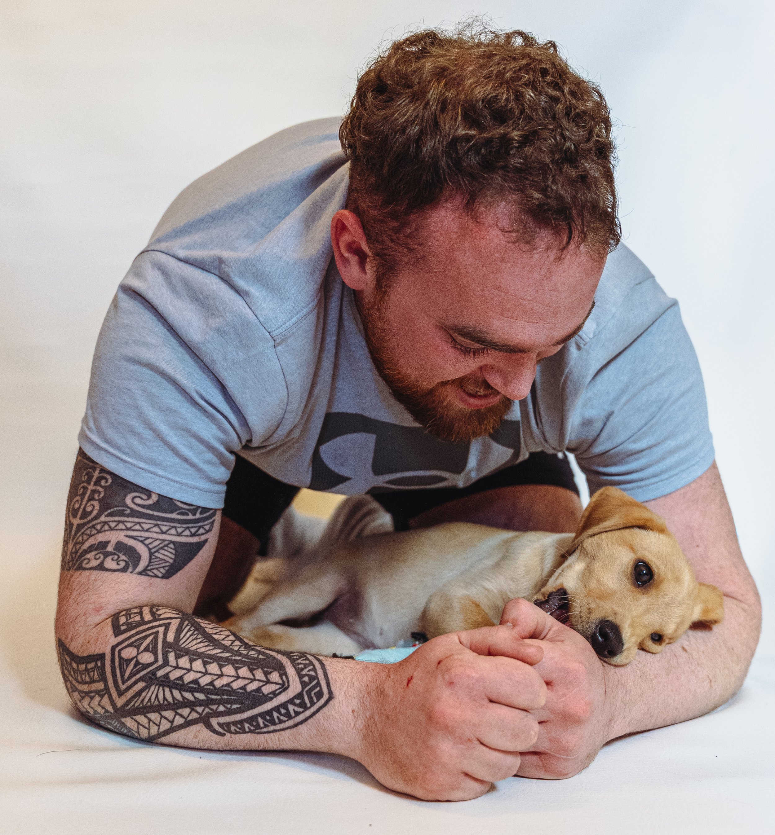 A man with curly hair and a tattooed arm is playing with a golden retriever puppy on a white surface.