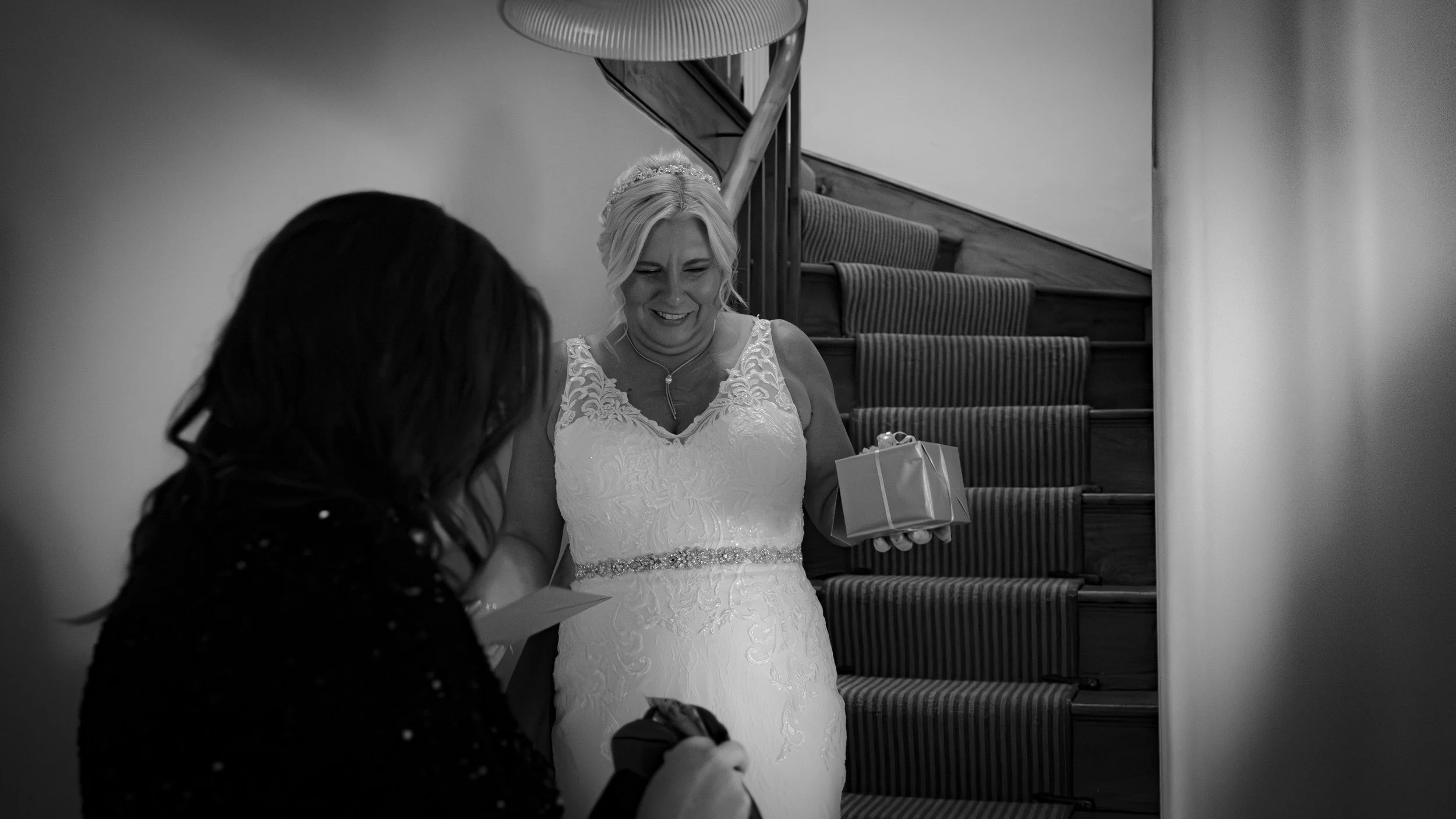 A bride in a white lace wedding gown receives a gift from a woman with dark hair, standing at the bottom of a staircase, with a smile.