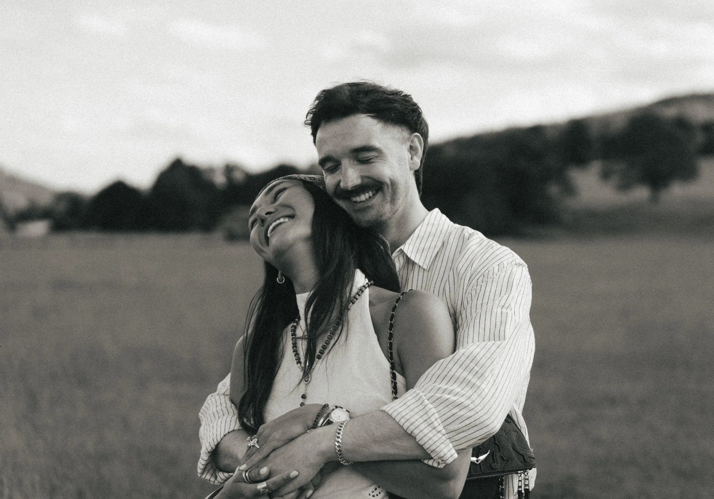 A black-and-white photo of a happy couple smiling and embracing outdoors, with a rural landscape and cloudy sky in the background.