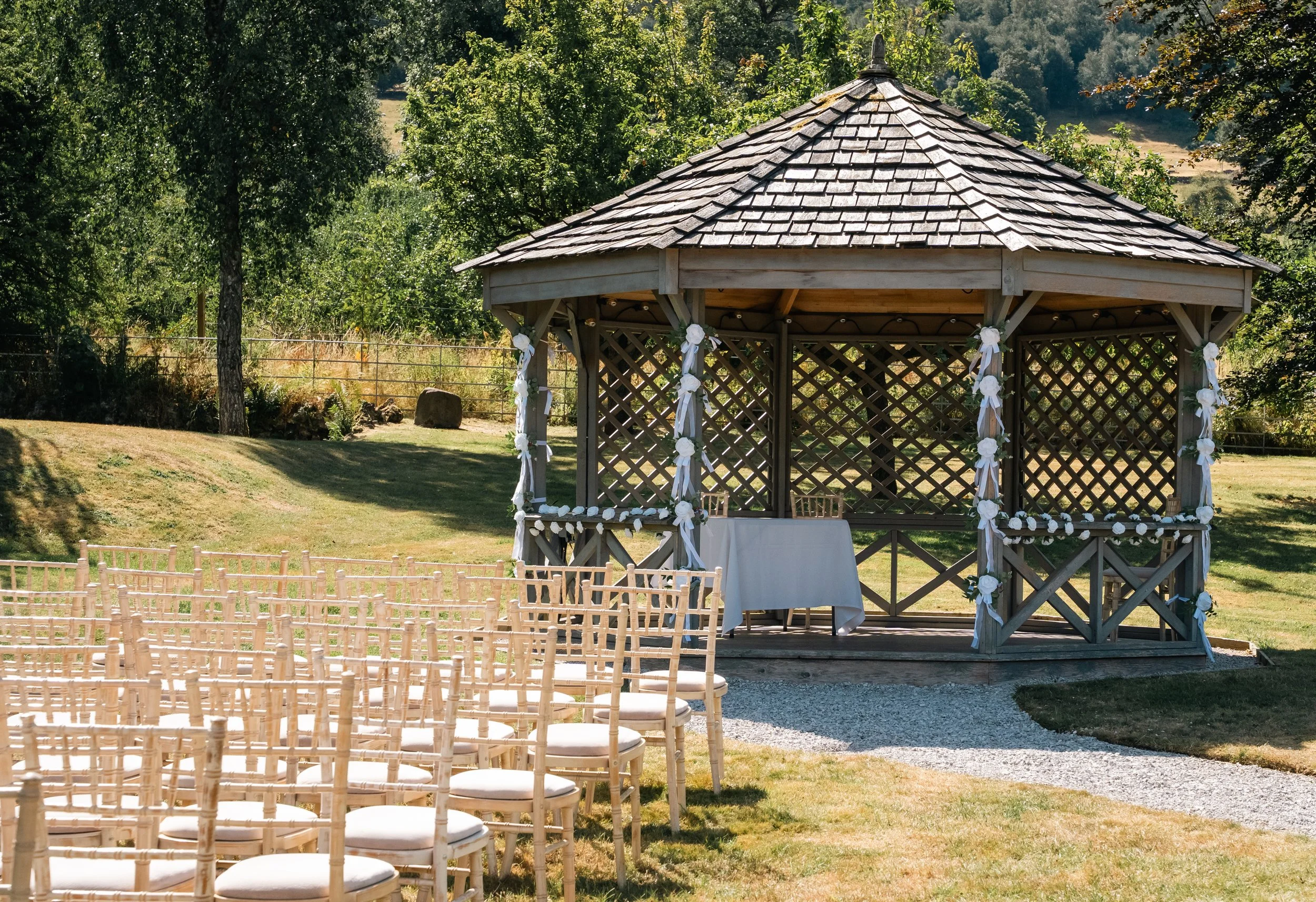 An outdoor wedding setup with rows of wooden chairs facing a wooden gazebo decorated with white ribbons and flowers, on a grassy area surrounded by trees, with a table underneath the gazebo.