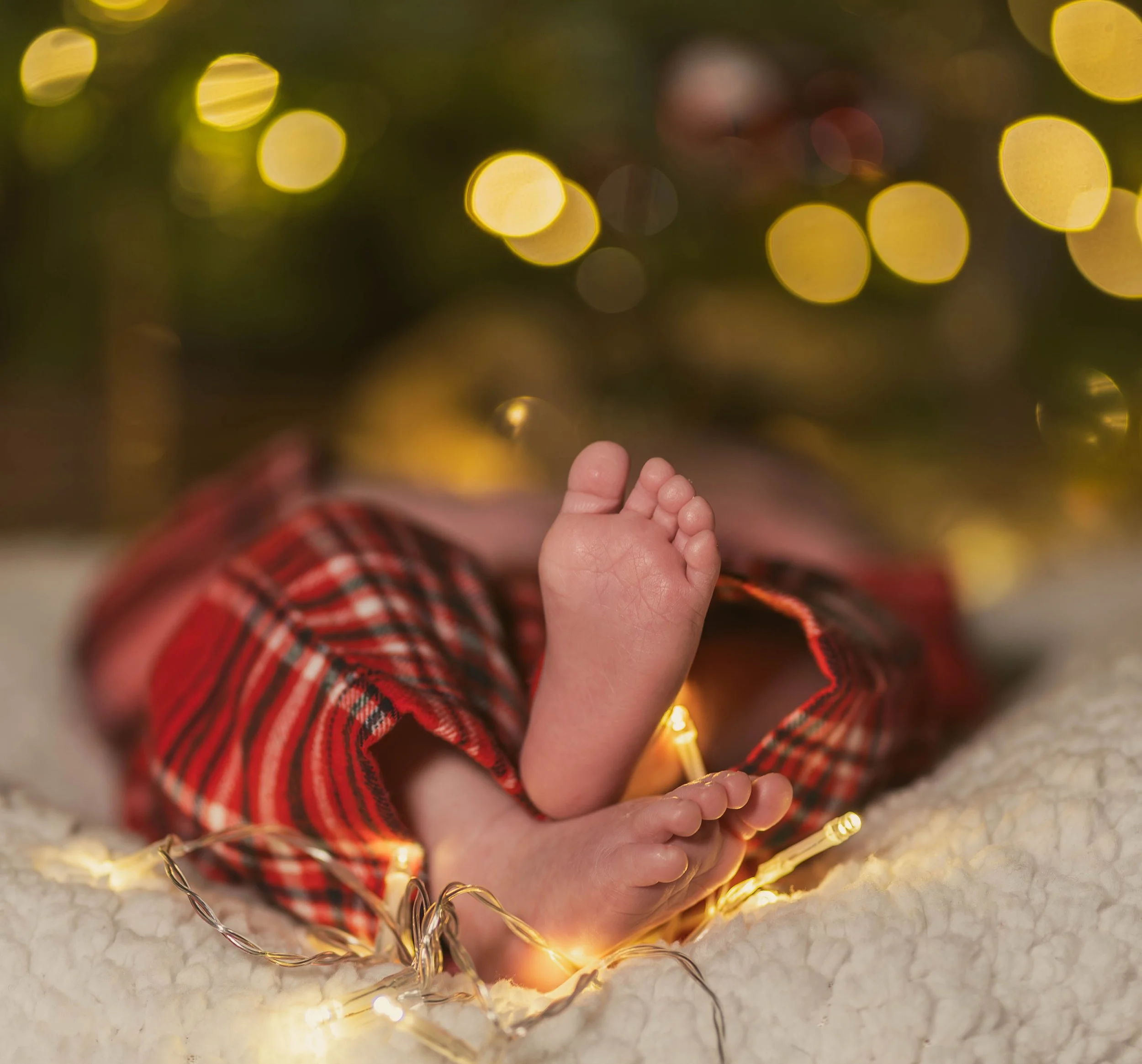 Close-up of baby feet resting on a soft, white blanket with Christmas lights and a blurred decorated Christmas tree in the background.