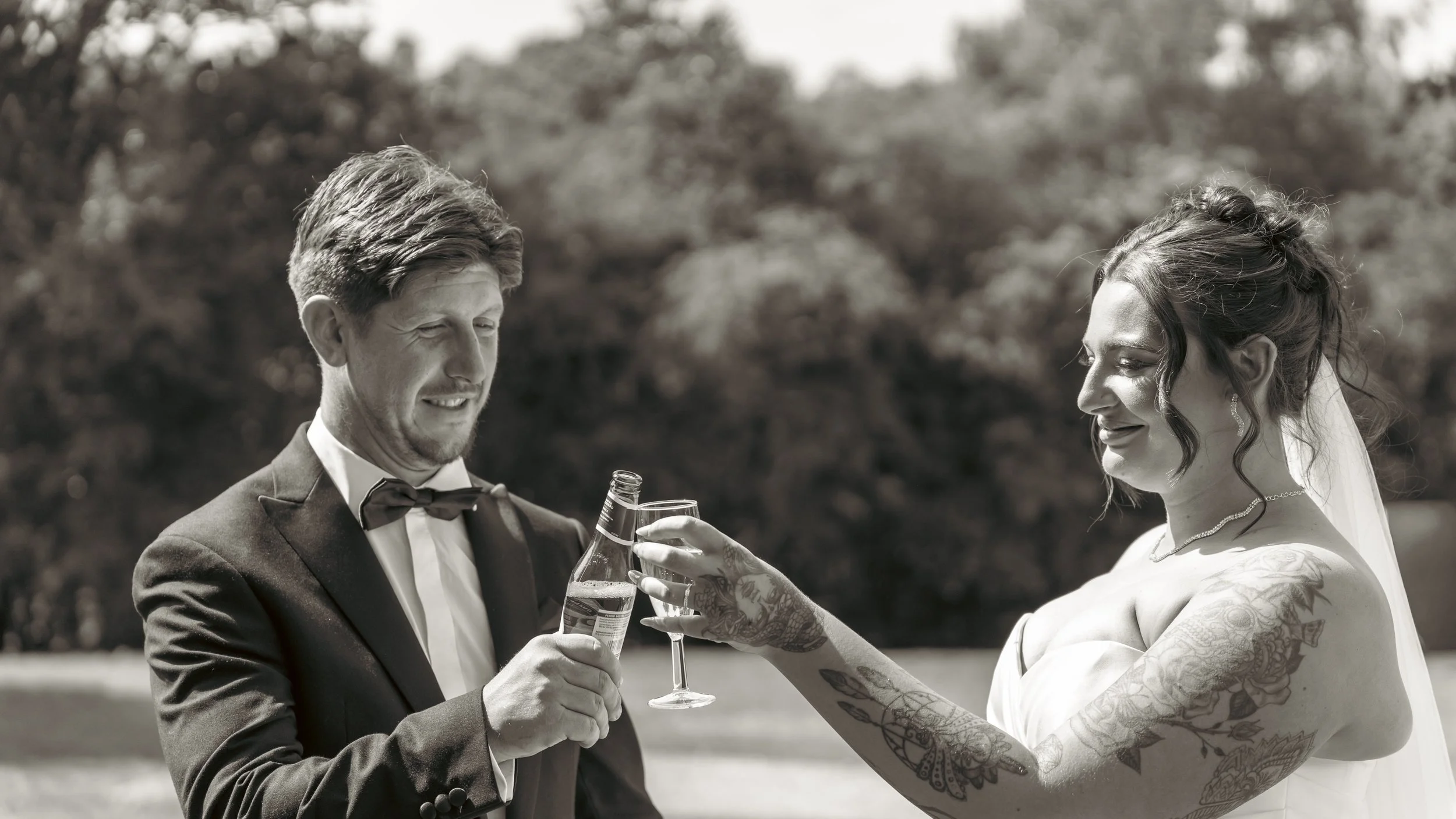 A black and white photo of a groom and bride outdoors, clinking beer bottles and glasses, smiling at each other. The groom is in a tuxedo with a bow tie, and the bride has tattoos on her arms and is wearing a wedding dress and jewelry.