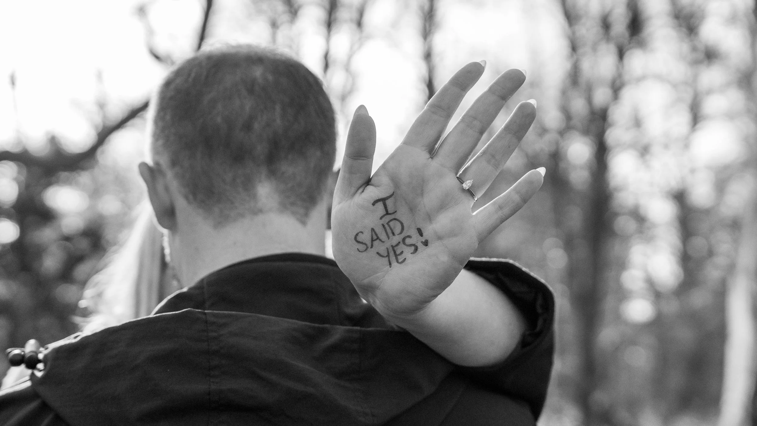 A person with short hair and a ring on their finger is holding up their hand, which has the words 'I SAID YES!' written on the palm, in front of their face, with trees in the background.