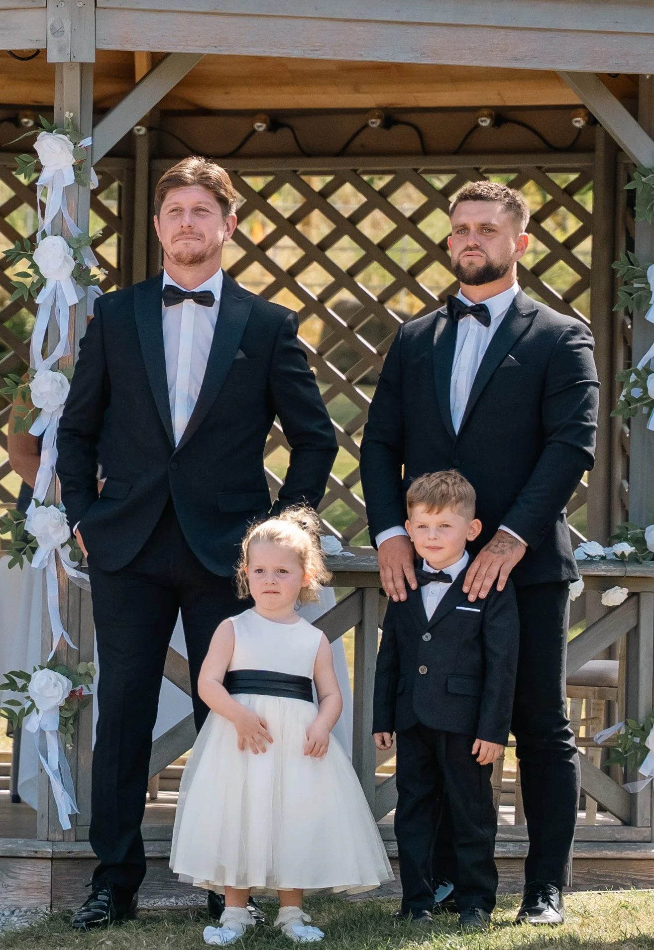 Two men in black tuxedos with bow ties standing on a wooden platform, surrounded by children in formal dresses and suits, at an outdoor wedding ceremony decorated with white flowers and greenery.