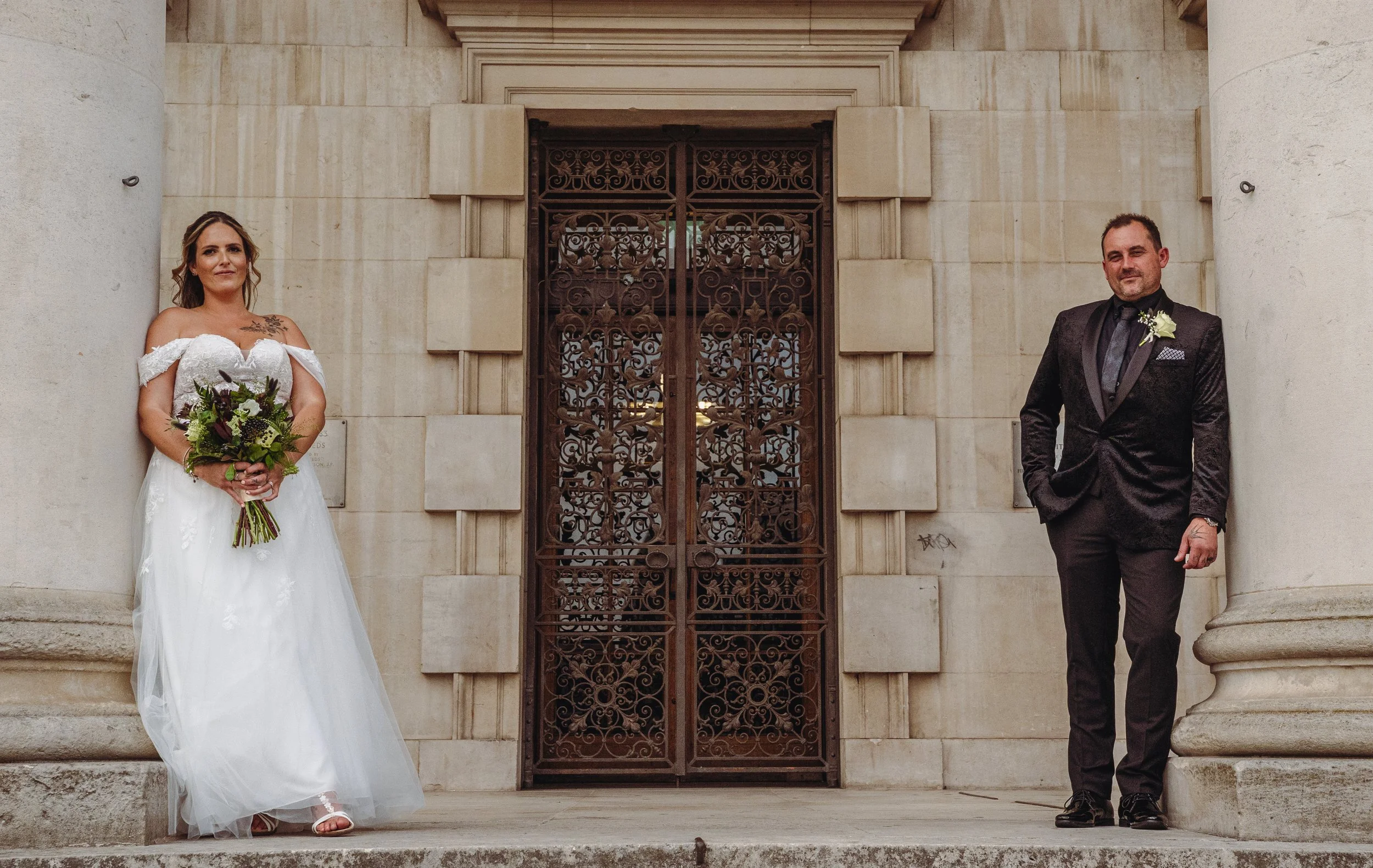 A bride in a white wedding gown holding a bouquet and a groom in a black suit standing on the steps outside a building with large stone columns and an ornate iron gate.