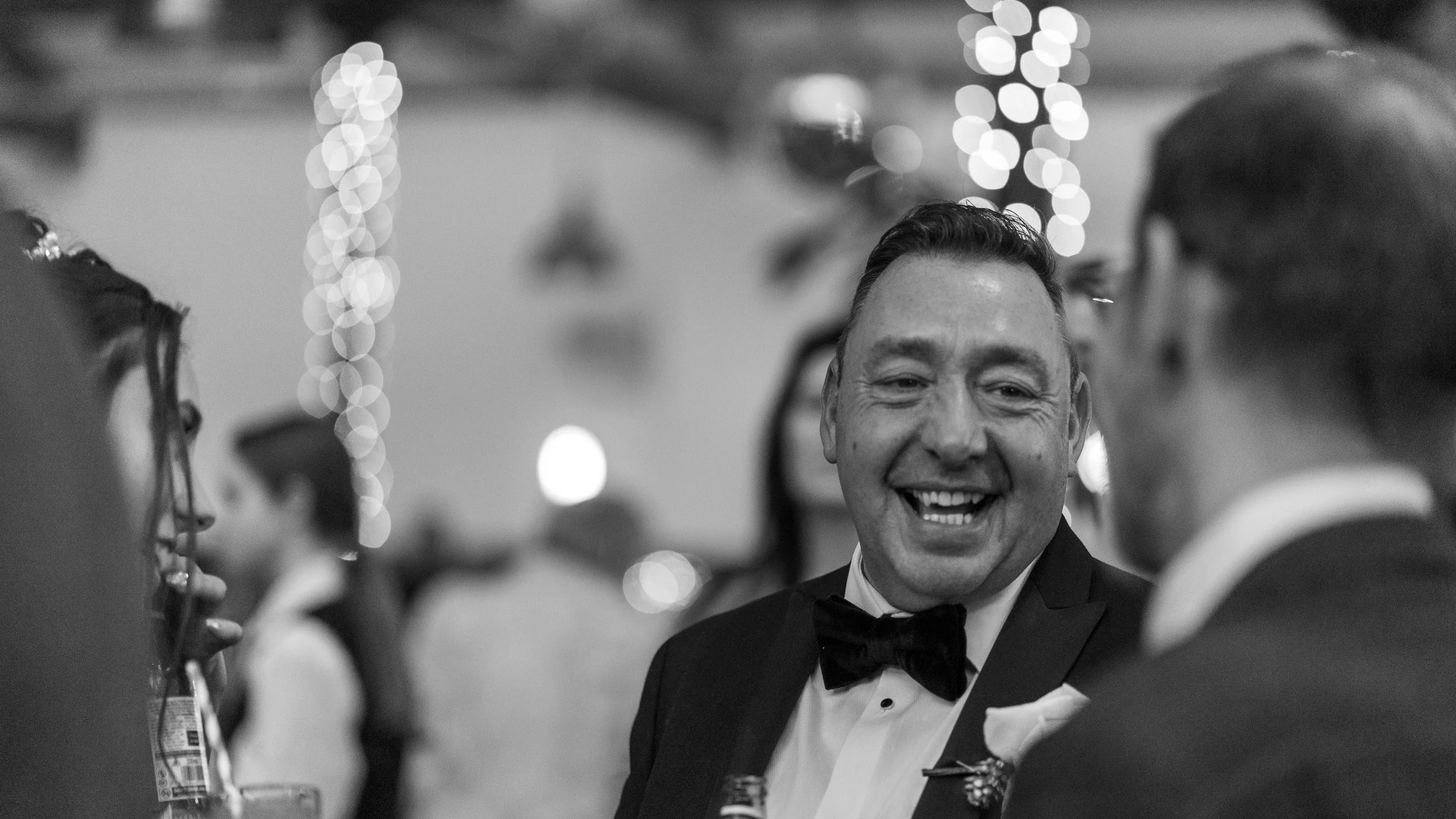 A group of people dressed in formal attire socializing at an event, with a man in a tuxedo smiling and talking to others in the foreground, decorated with string lights in the background.