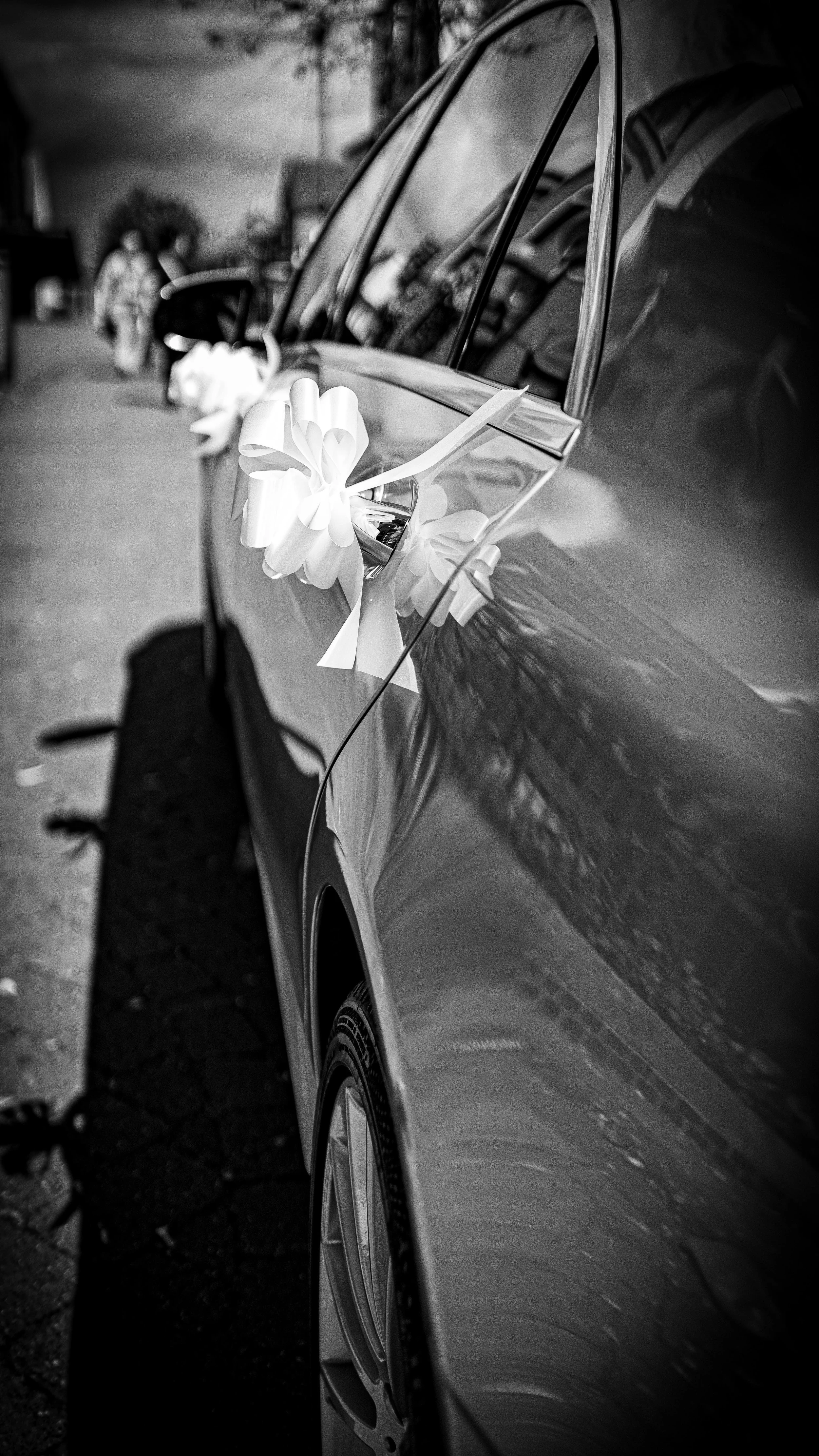 A car decorated with a white ribbon and bow for a wedding, parked outside on a street.