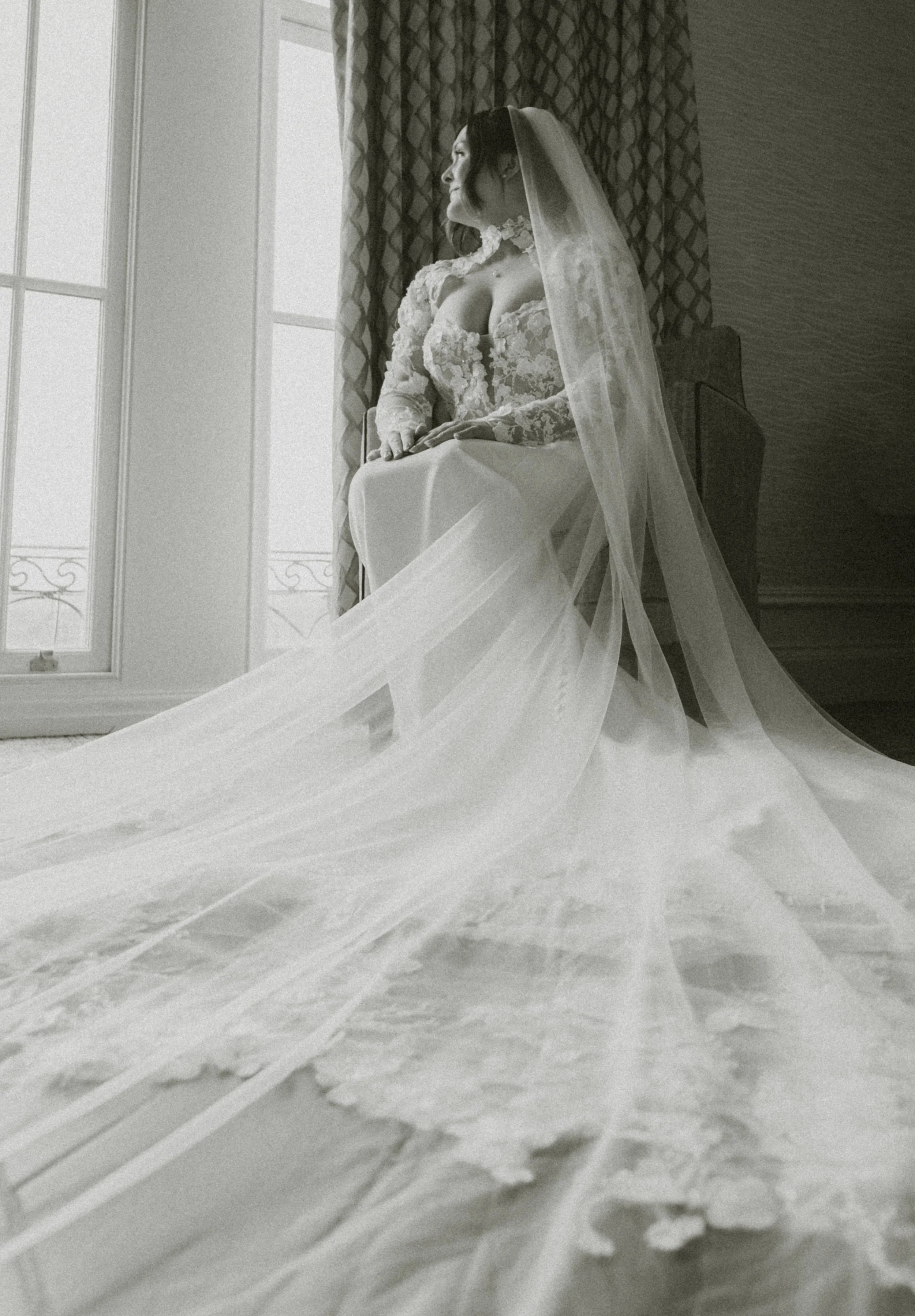 A woman dressed as a bride in a lace wedding dress with a veil, sitting by a window with patterned curtains, looking outside.