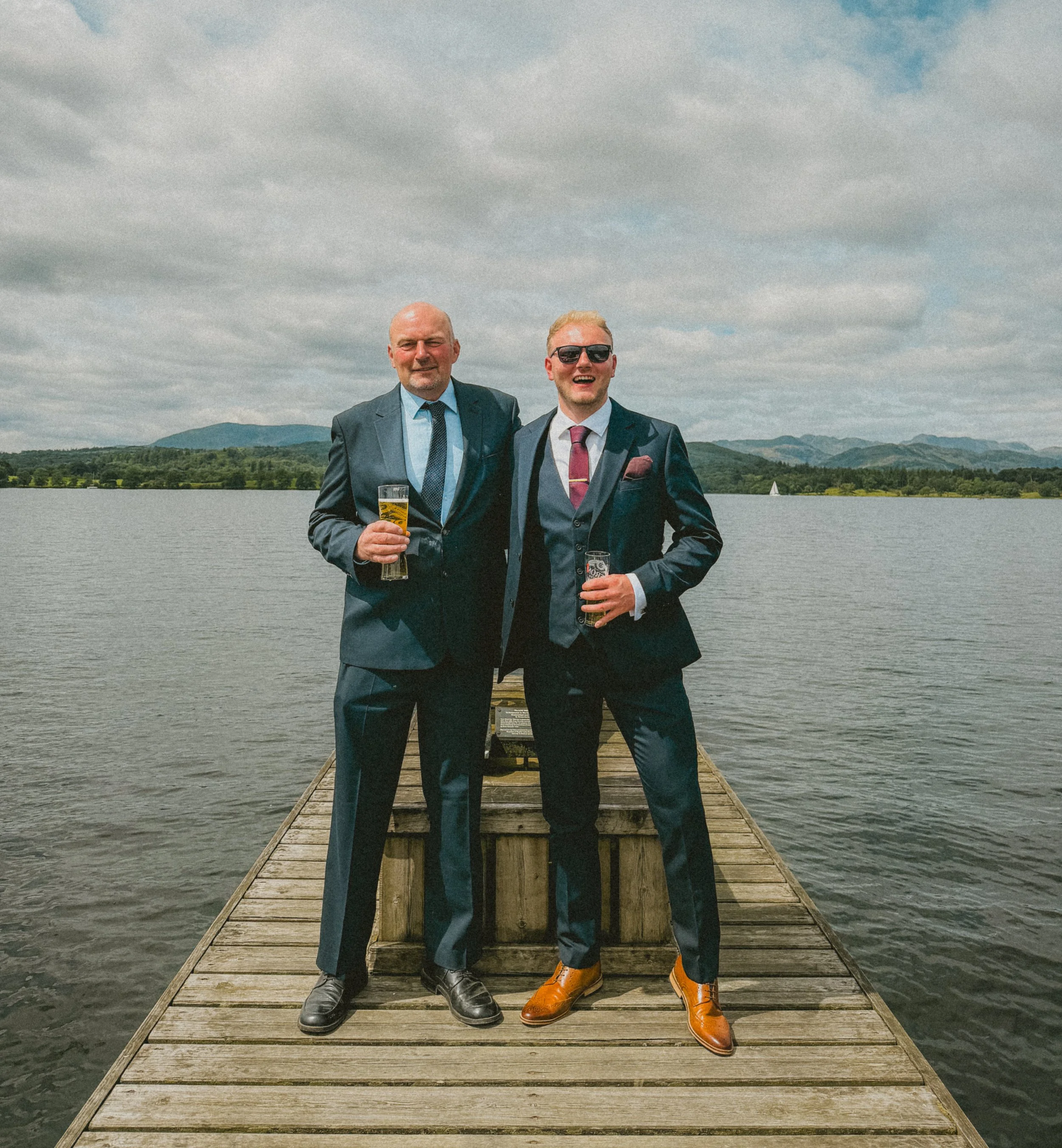 Two men in suits standing on a wooden dock by a lake, holding glasses of beer, with mountains and cloudy sky in the background.