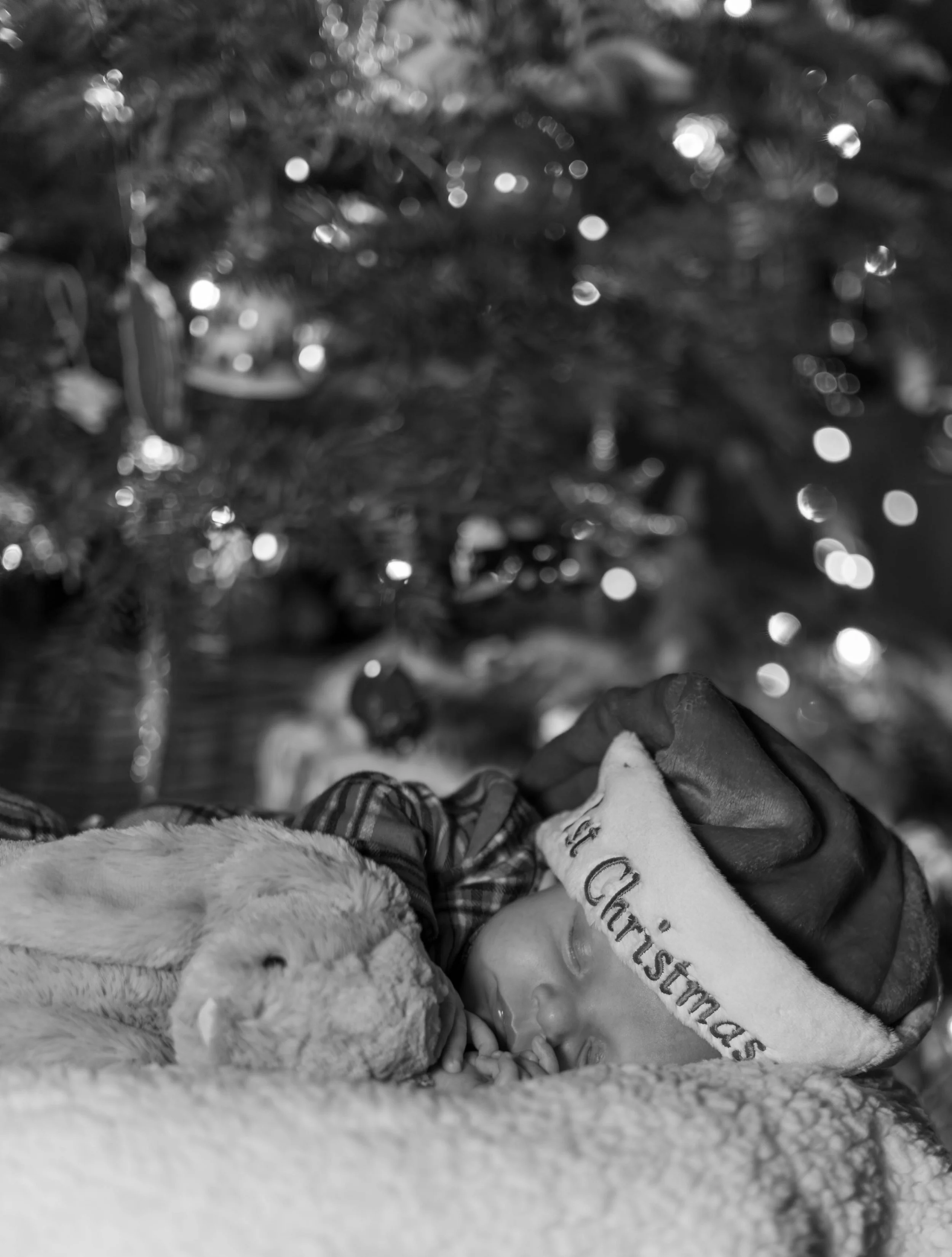 A sleeping baby wearing a Christmas hat with the words ‘Merry Christmas’, resting with a stuffed animal, in front of a blurred decorated Christmas tree.