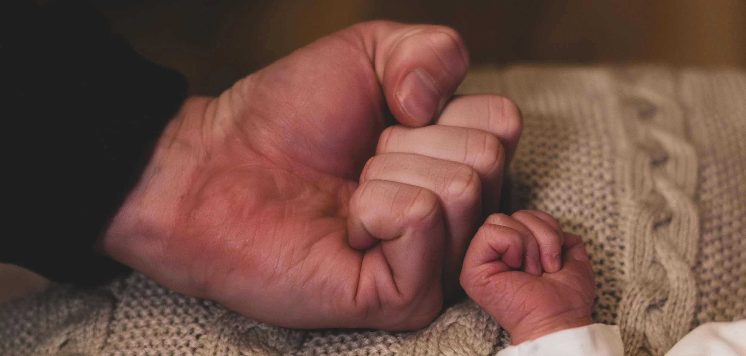Close-up of a large adult hand gently holding a tiny baby's hand, resting on a knitted blanket.