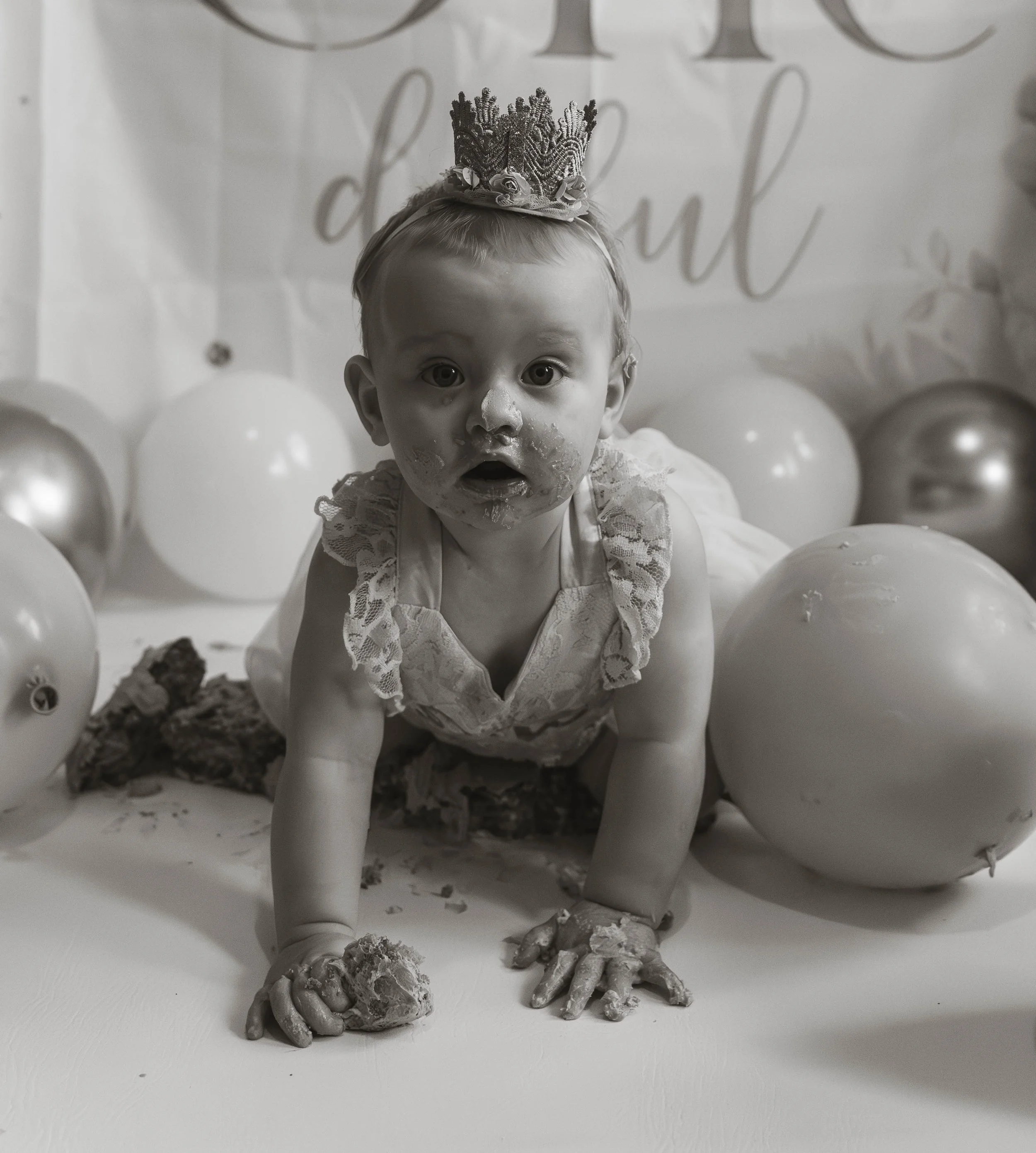 A black-and-white photo of a young child with a crown on her head, surrounded by balloons, with her face and hands covered in cake or frosting.