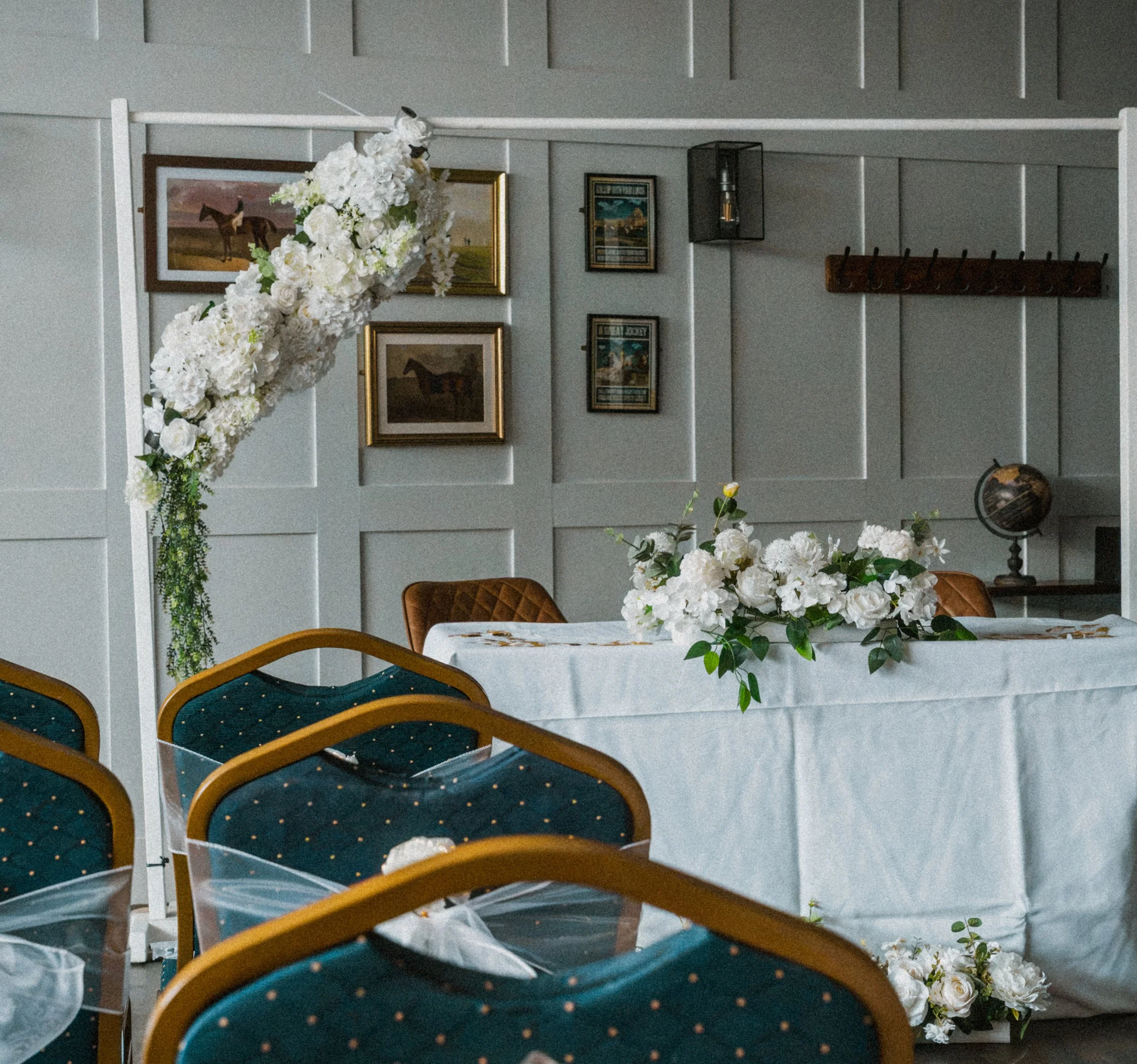 Interior of a room decorated with white paneled walls, framed artwork, and hanging black light fixtures. A cluster of artificial white flowers and green foliage decorates a table, and green plants hang on the wall. Several navy blue chairs with woode