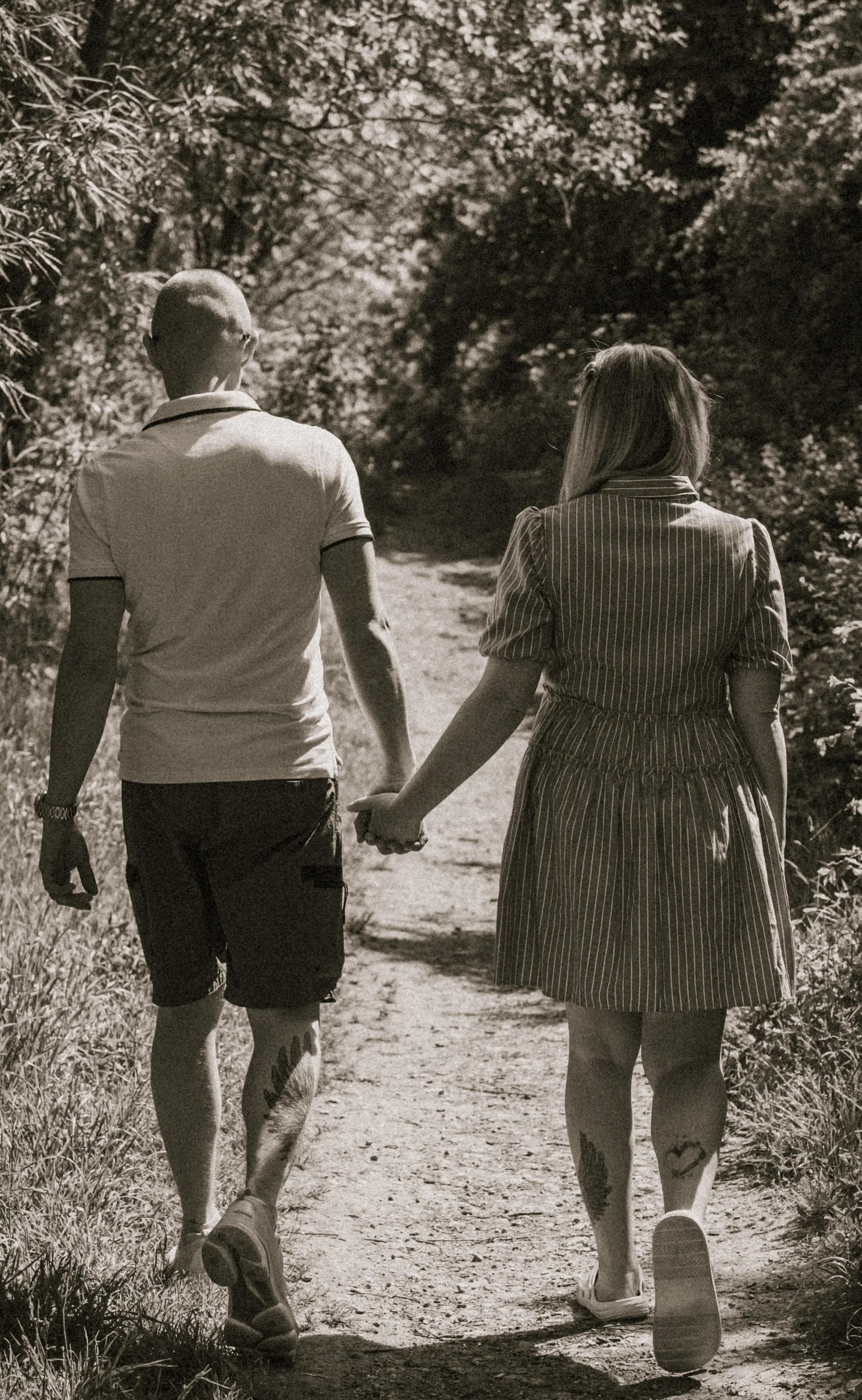 A black and white photo of a couple walking hand-in-hand on a dirt trail surrounded by trees. The man has a shaved head, sunglasses, a polo shirt, shorts, and tattoos on his legs. The woman has long hair, a striped dress, and tattoos on her legs. The