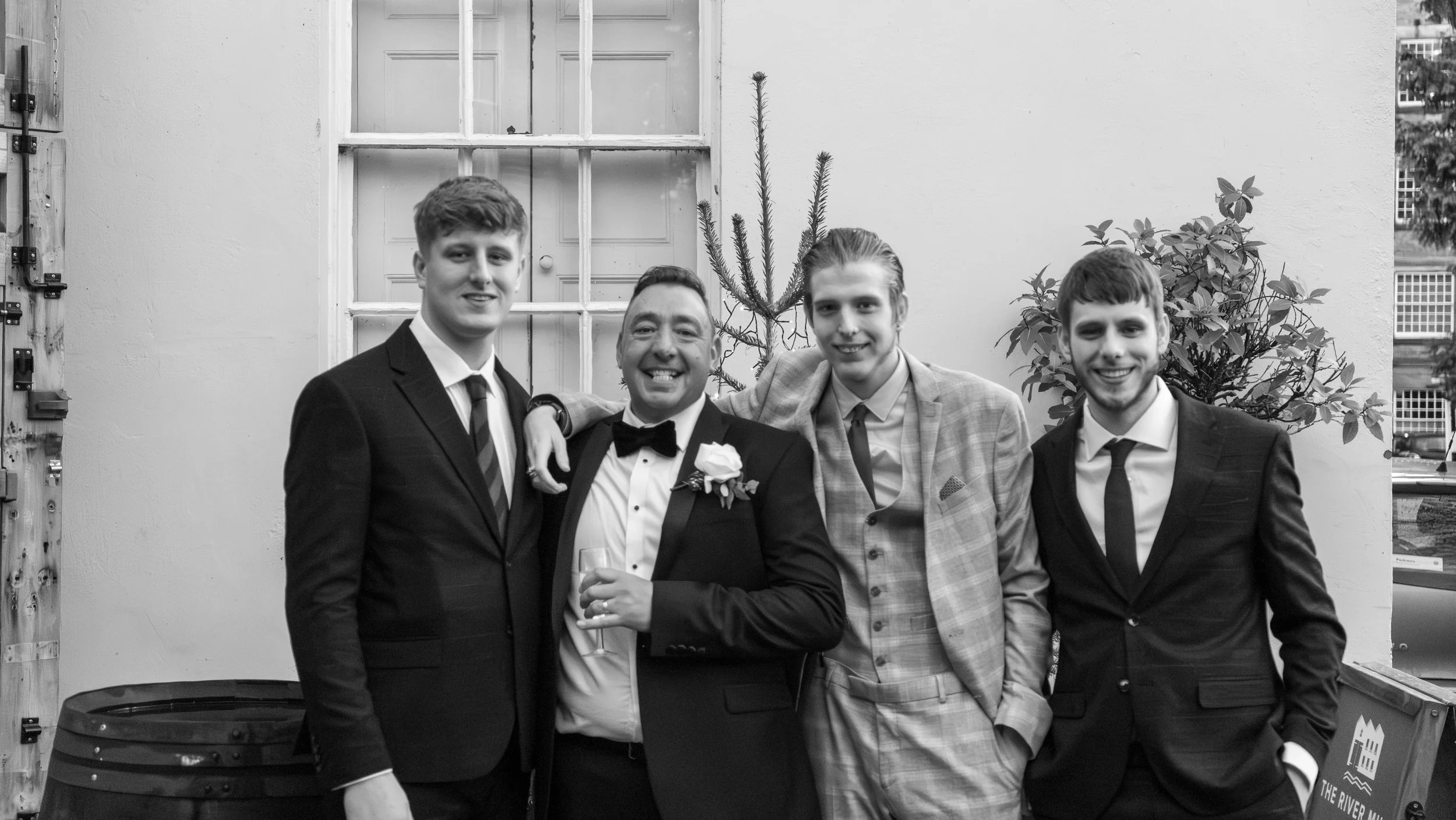 Four men dressed in formal suits, three younger men and one in a tuxedo with a boutonniere, standing outdoors against a wall with a window, plants, and a small tree behind them, smiling for the photo.