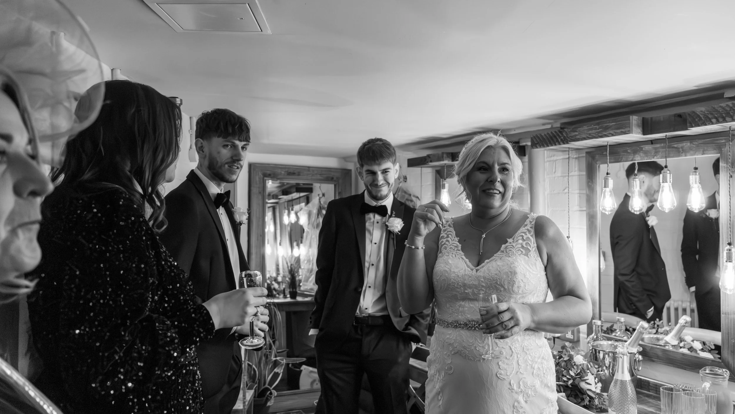 Black and white photo of a wedding reception scene with a woman in a white lace wedding dress smiling and holding a glass, surrounded by people in formal attire, with hanging light bulbs and a mirror in the background.
