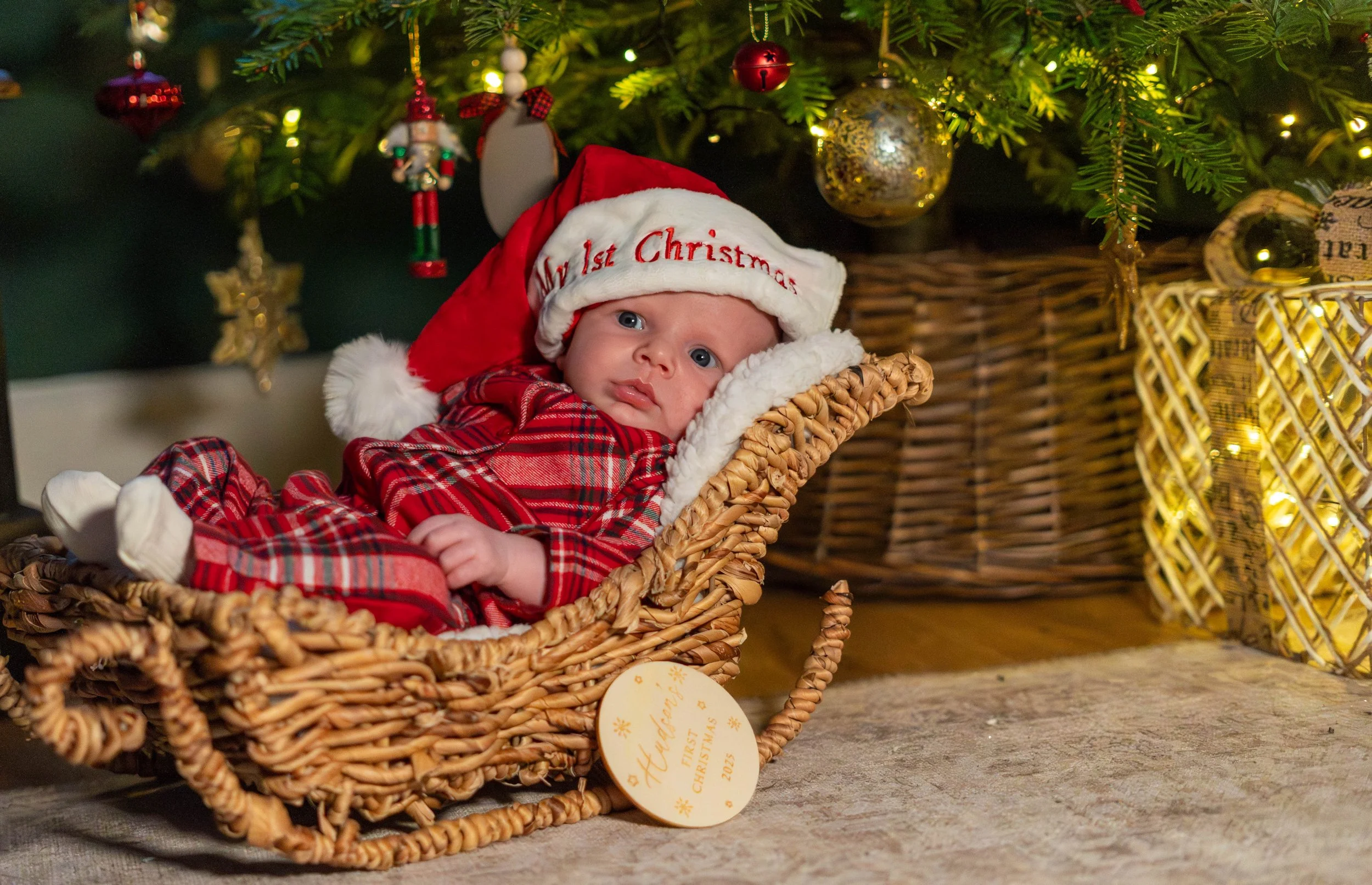 A baby dressed in red plaid pajamas and a Santa hat that says "My 1st Christmas" is lying in a wicker basket under a decorated Christmas tree with lights and ornaments.