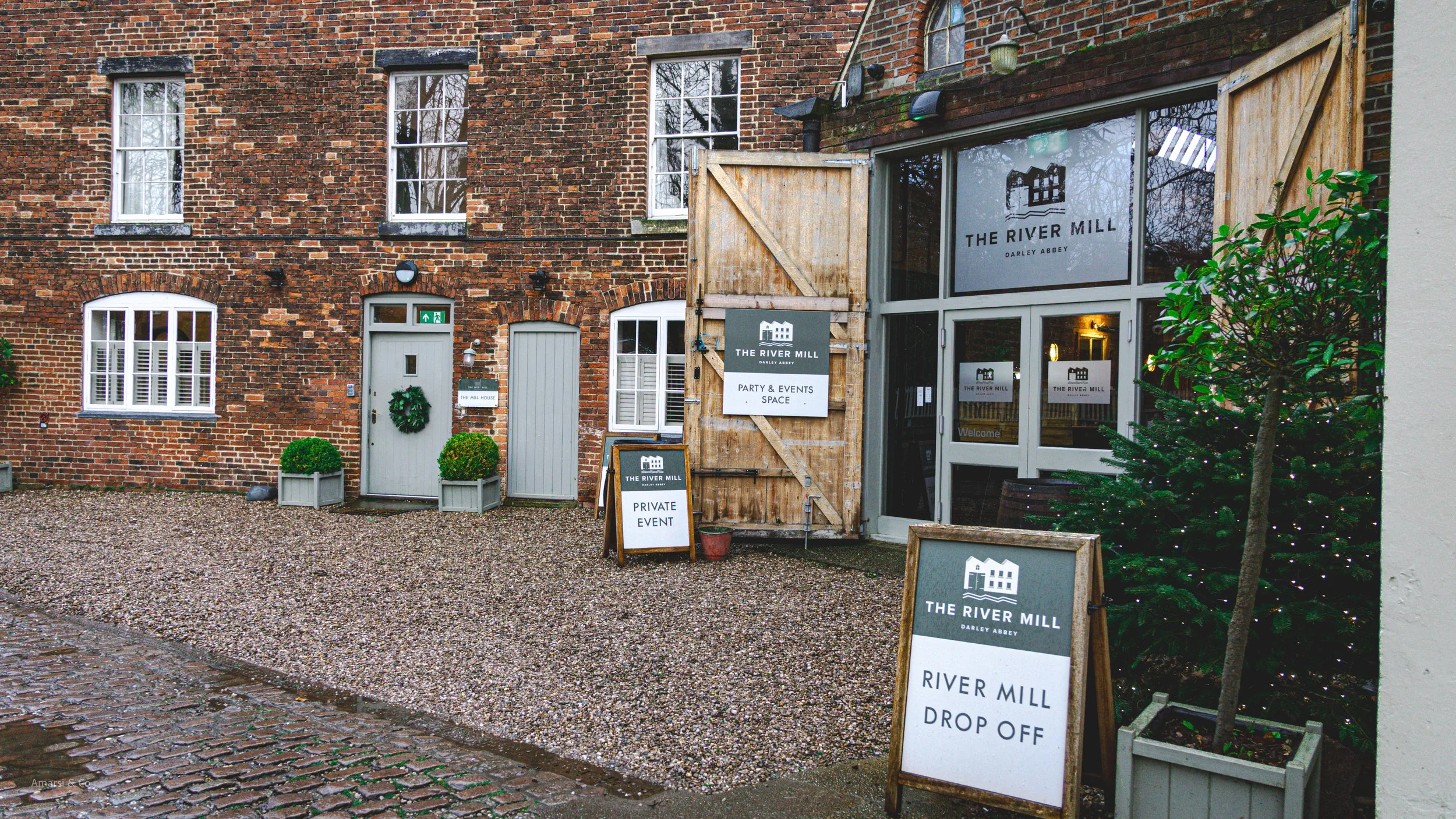 Exterior view of The River Mill, a brick building with white framed windows. There are signs indicating it is a party and events space, with a private event notice and a drop-off area. A Christmas tree decorated with lights is visible on the right, a