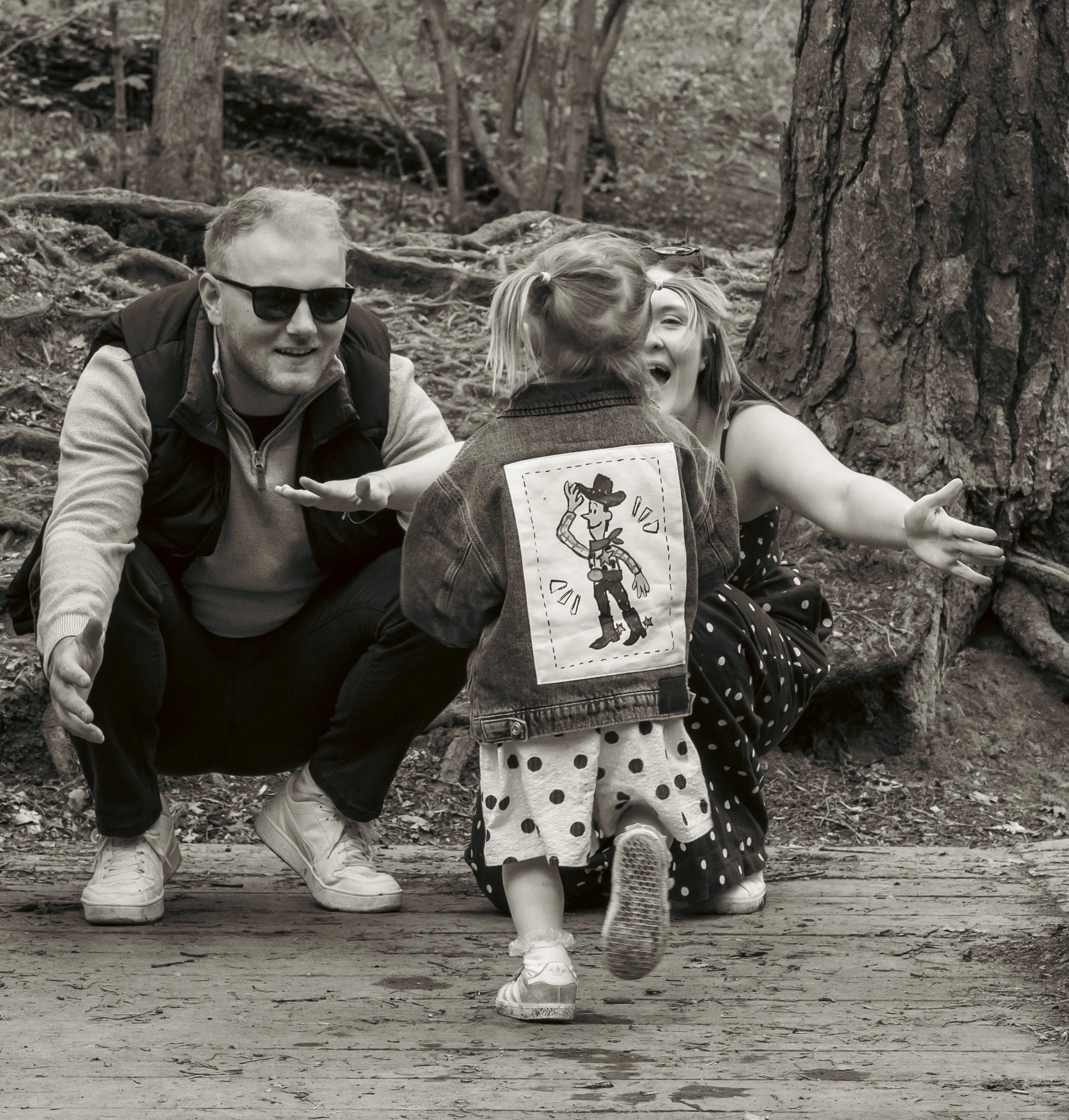 Three children and a man playing outdoors in a wooded area. The children are running and laughing, with one girl wearing polka dot skirt and Mickey Mouse shirt, and the man wearing sunglasses and a jacket. The background shows trees and roots.