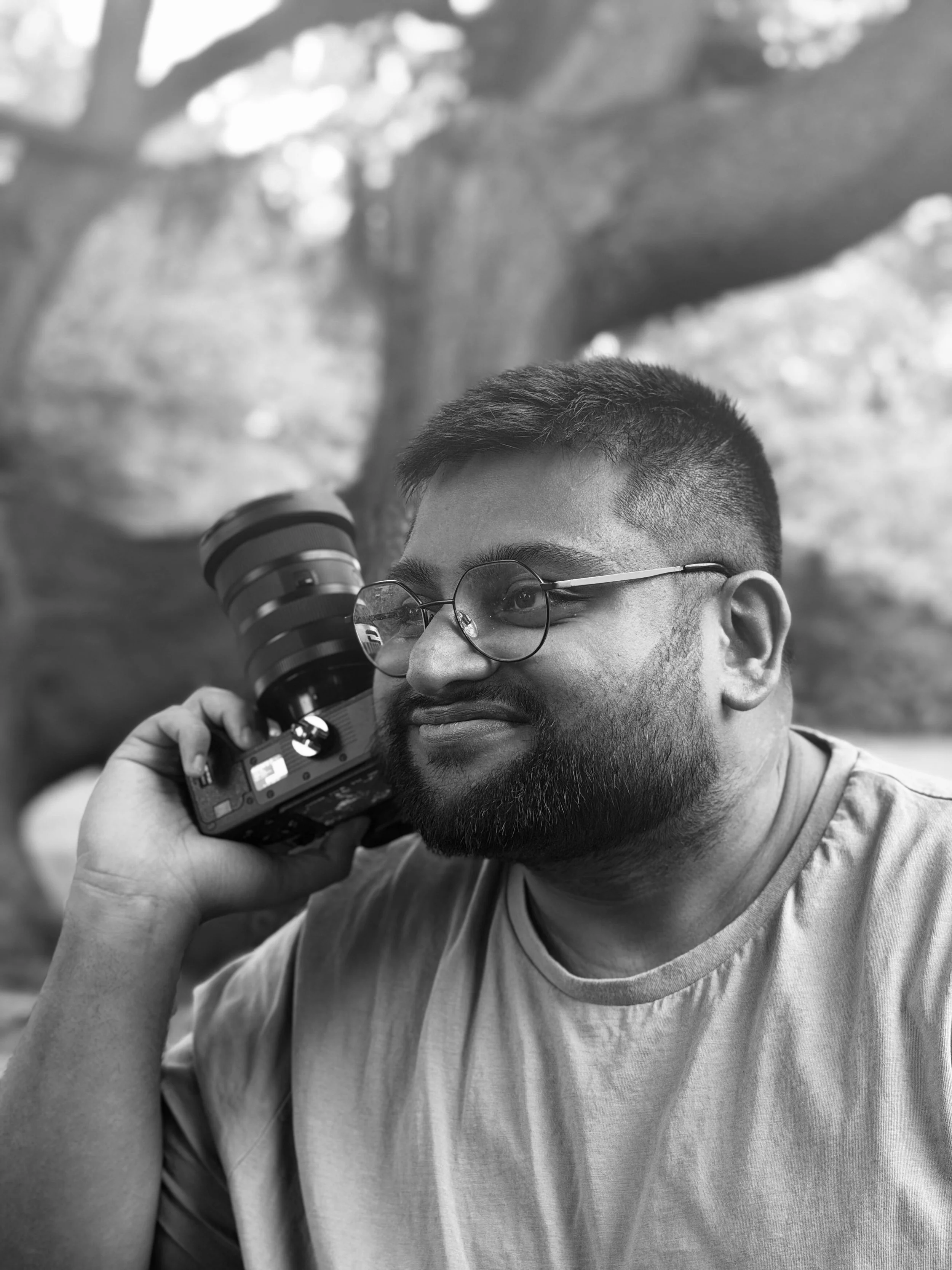 A man with glasses and short hair is holding a camera to his face, appearing to be taking a photo outdoors. The background includes blurred trees and foliage.