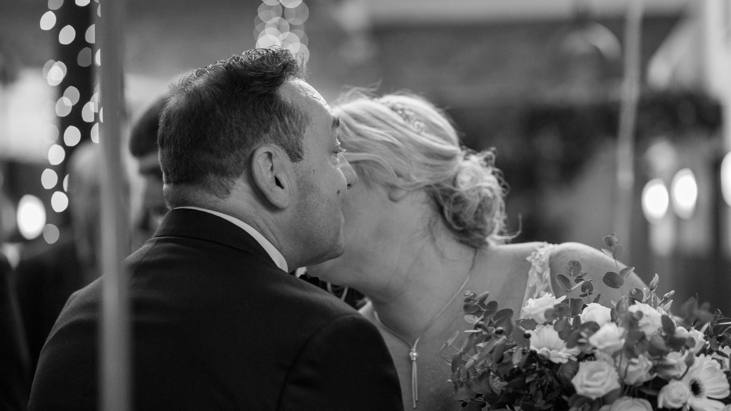 A black and white photo of a man and woman sharing a kiss; the woman holds a bouquet of flowers.