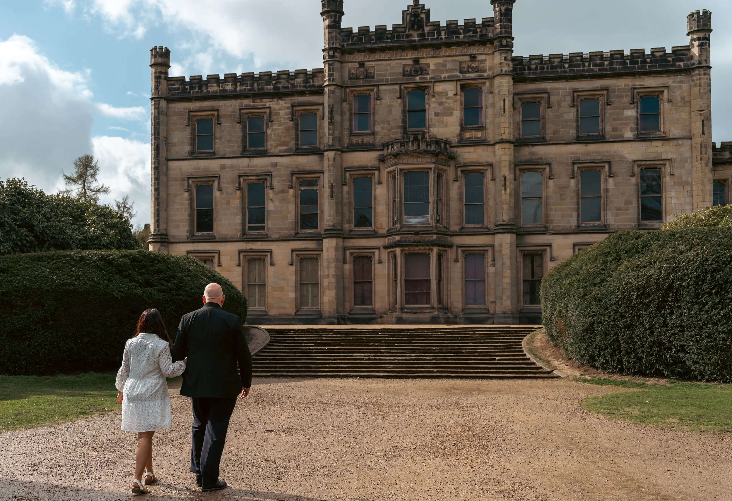 Couple walking up steps toward a historic stone building on a cloudy day, with lush bushes on either side.