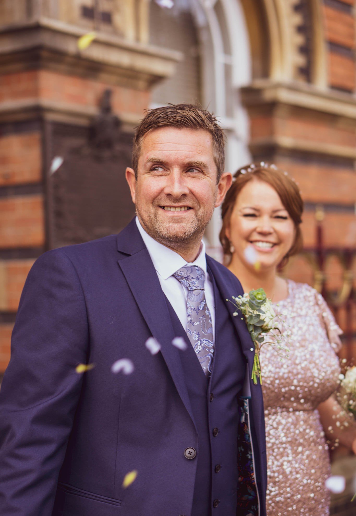 A groom in a navy suit with a floral tie and a boutonniere, smiling as guests celebrate at a wedding outside a brick building.