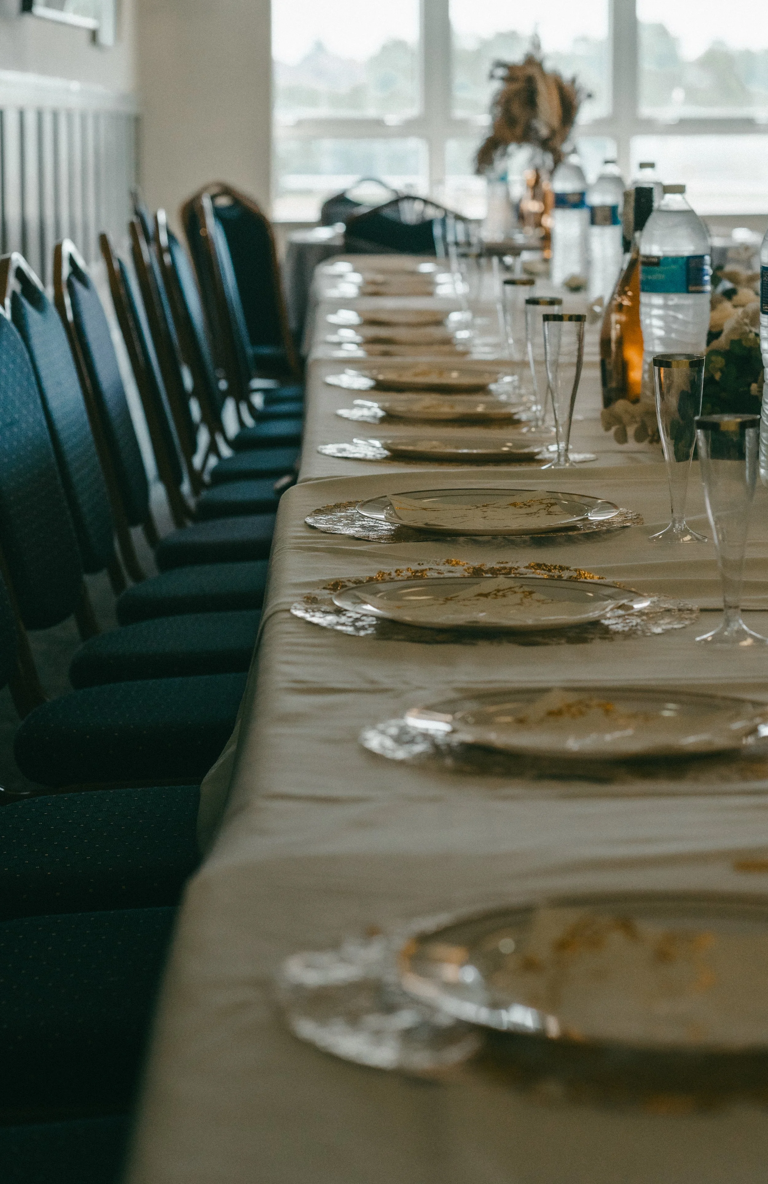 A long banquet table set for a meal with plates, napkins, and glasses, surrounded by chairs, with water bottles and some decorative items in the background.