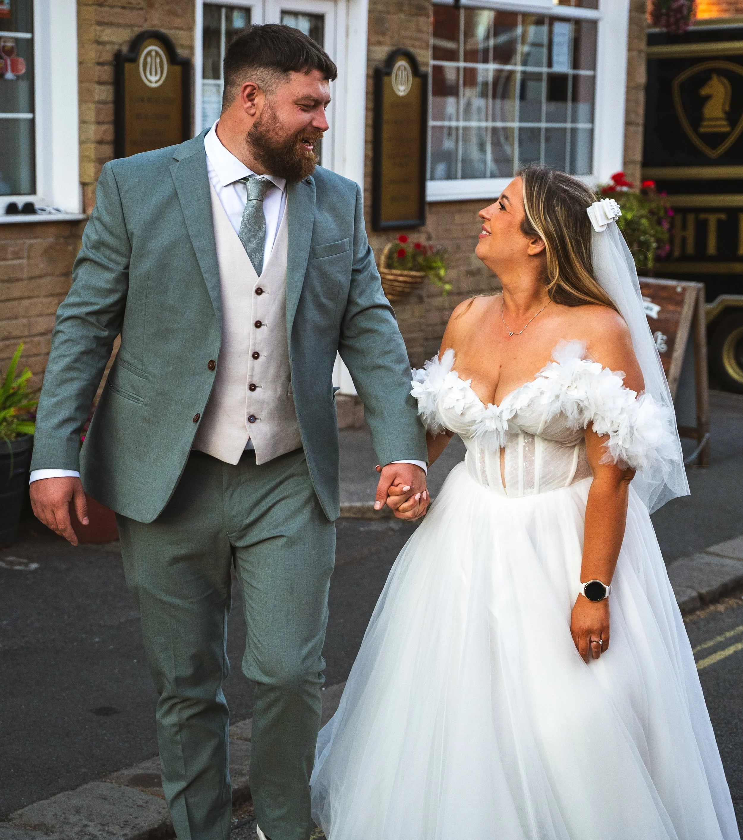 A man and woman holding hands, smiling at each other outside a building, possibly during a wedding or special event.