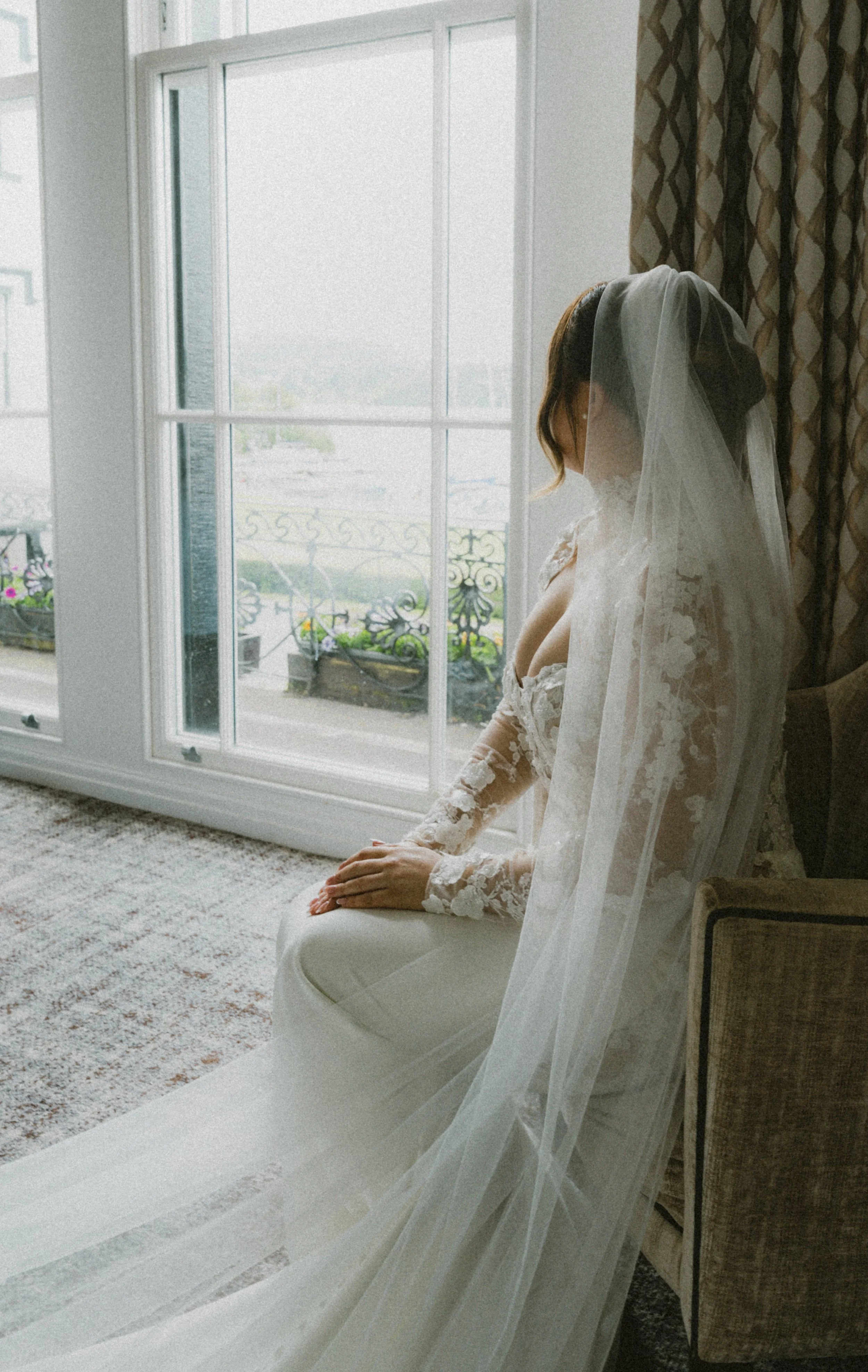 A bride in a lace wedding gown and veil sitting on a chair by a large window with curtains, looking down, with flowers and a balcony visible outside.