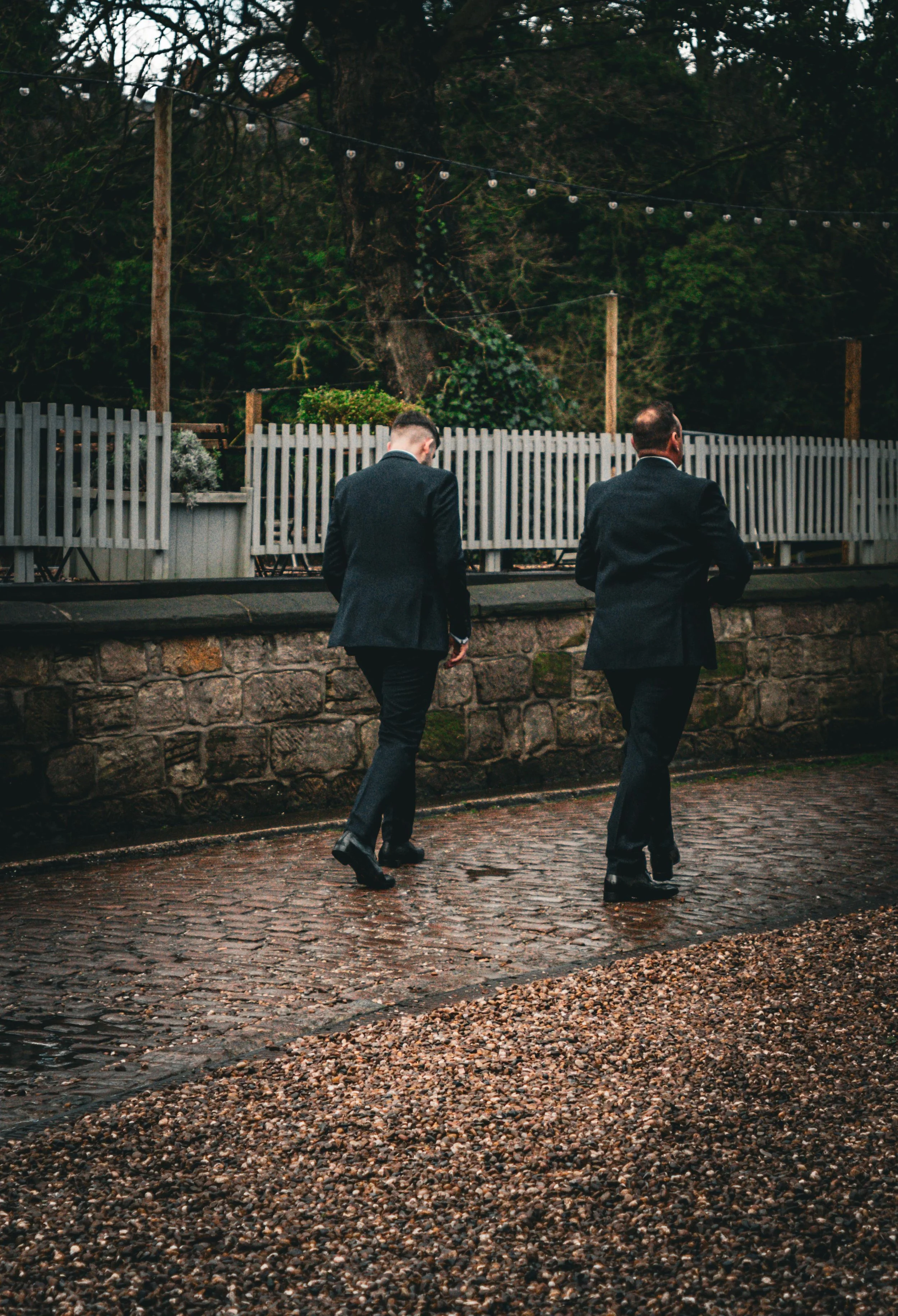 Two men in black suits walking on wet cobblestone street near a stone wall, with trees and string lights in the background.