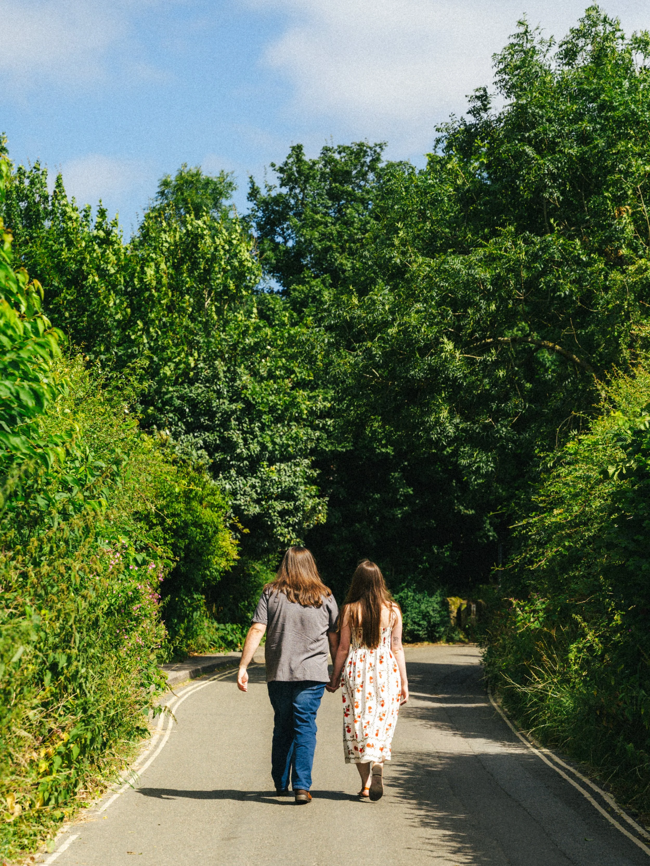 Two women walking down a curved road surrounded by lush green trees, with a blue sky overhead.