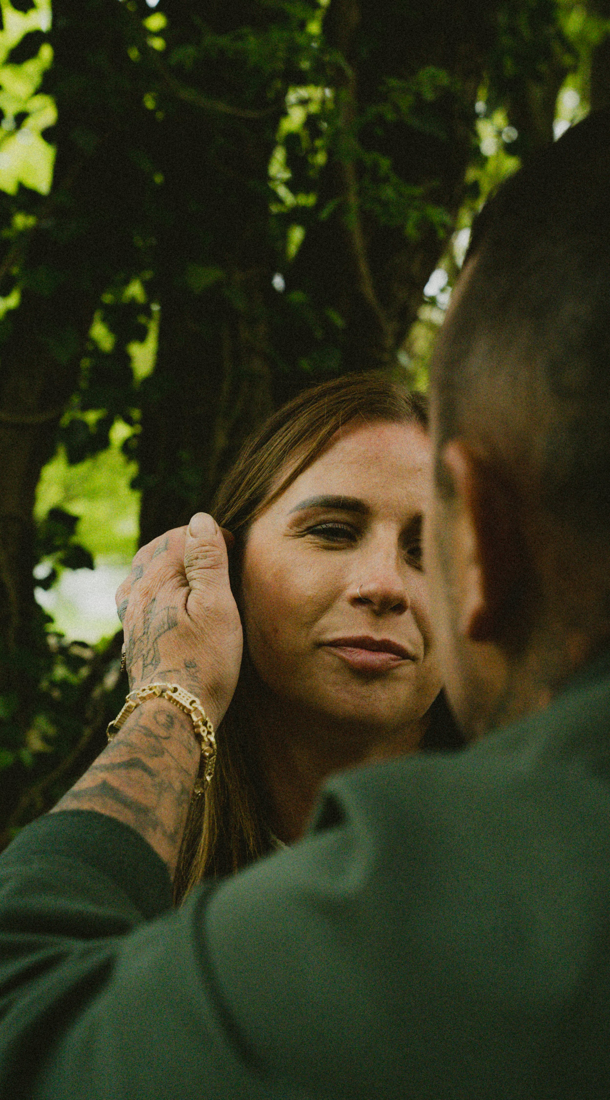 A woman with makeup and a nose ring looks at a person in front of her as they adjust her hair or face. The scene is outdoors with green foliage in the background.
