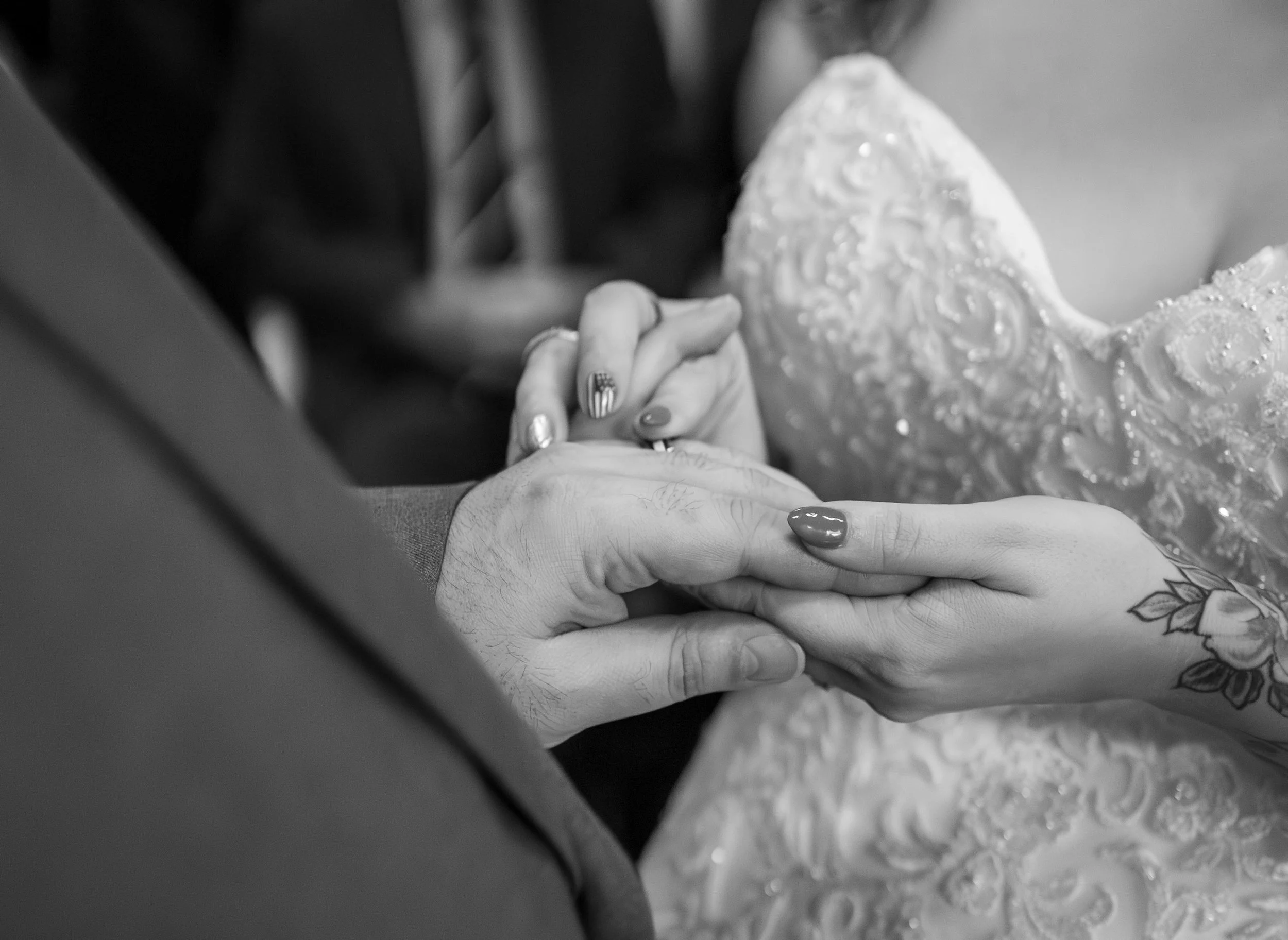 Close-up of a couple holding hands during a wedding ceremony, with the bride's hand resting on top of the groom's hand. The bride's dress has intricate embroidery, and she has tattoos and painted nails.