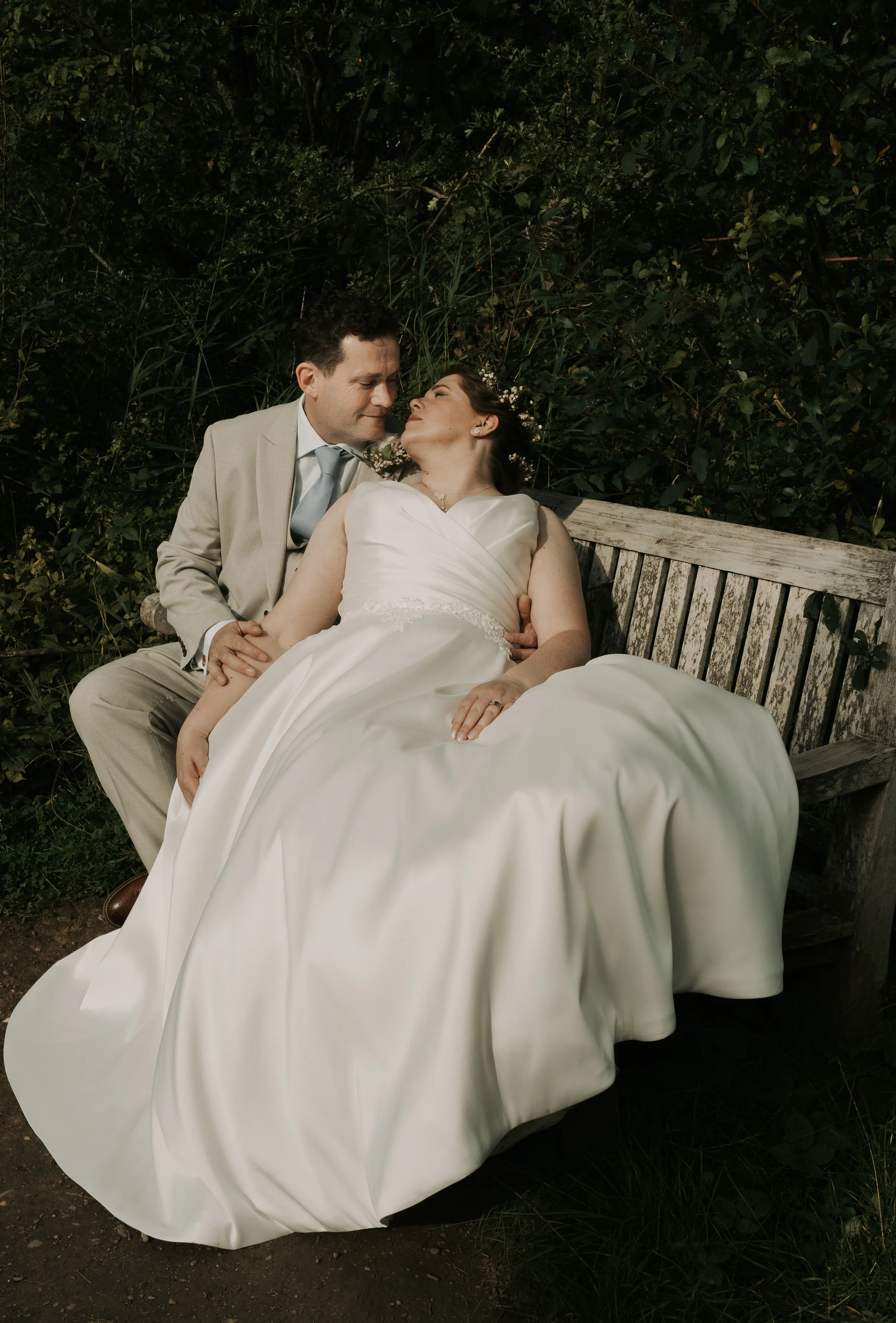 A newlywed couple sitting on a wooden bench outdoors surrounded by greenery, At The Glade, Rosliston