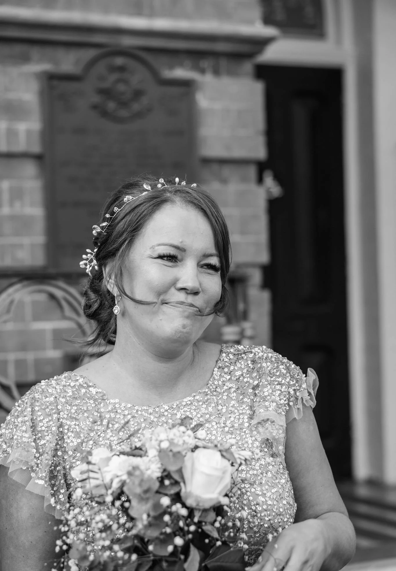 A smiling woman wearing a sequined dress and a floral headband, holding a bouquet of flowers, standing in front of a brick building.
