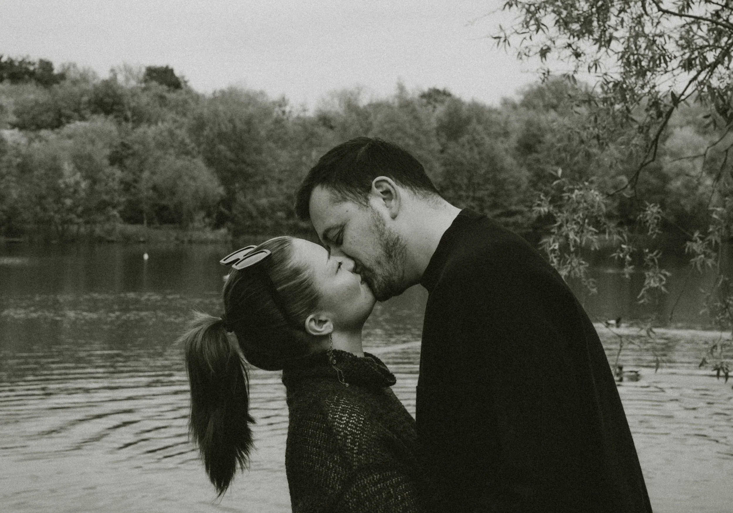 A couple sharing a kiss outdoors near a body of water, with trees and foliage in the background.