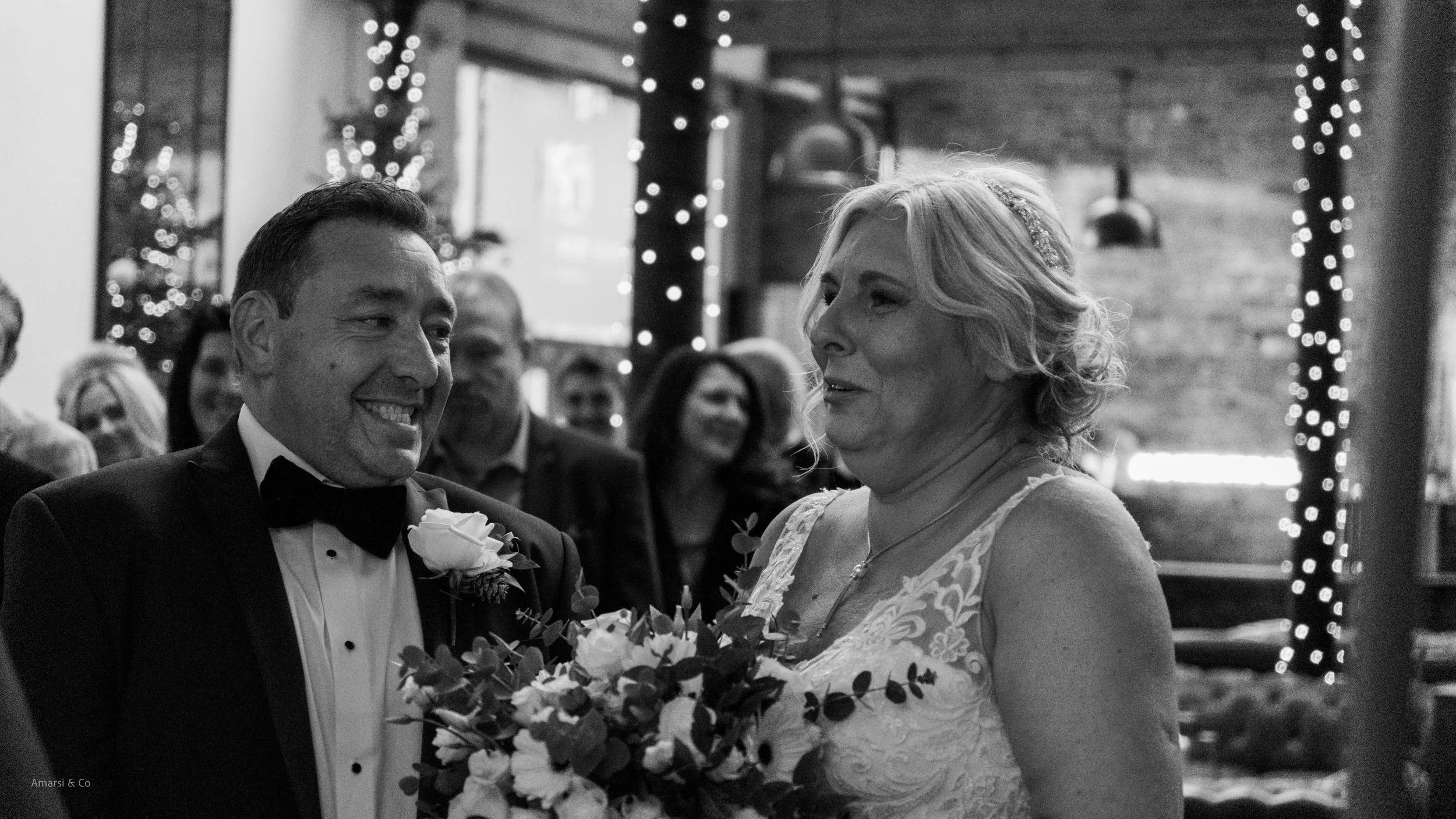 A black and white photo of a wedding scene with a groom in a tuxedo and a bride in a lace wedding dress, smiling at each other. The bride is holding a bouquet of flowers, and there are guests in the background with string lights and decorations.