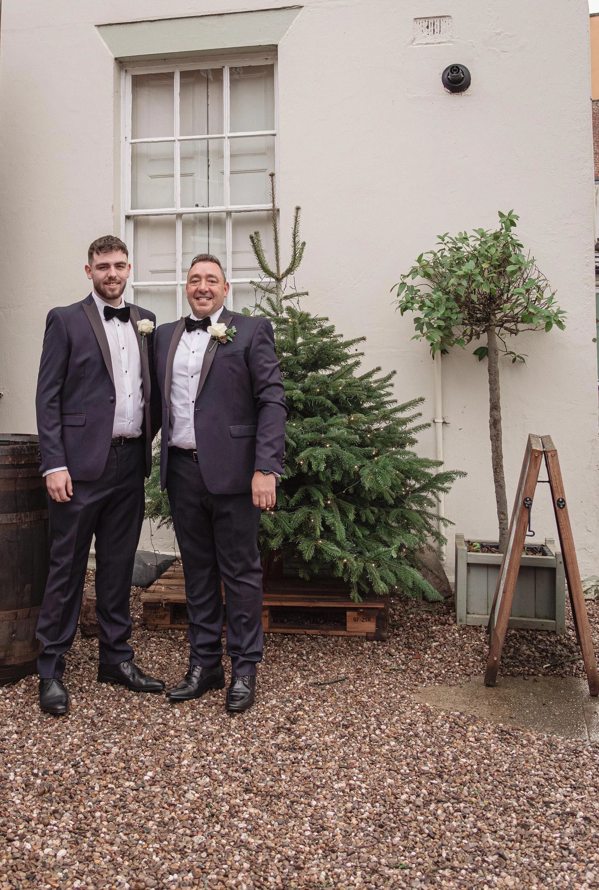 Two men in tuxedos standing outdoors beside a decorated Christmas tree, smiling.