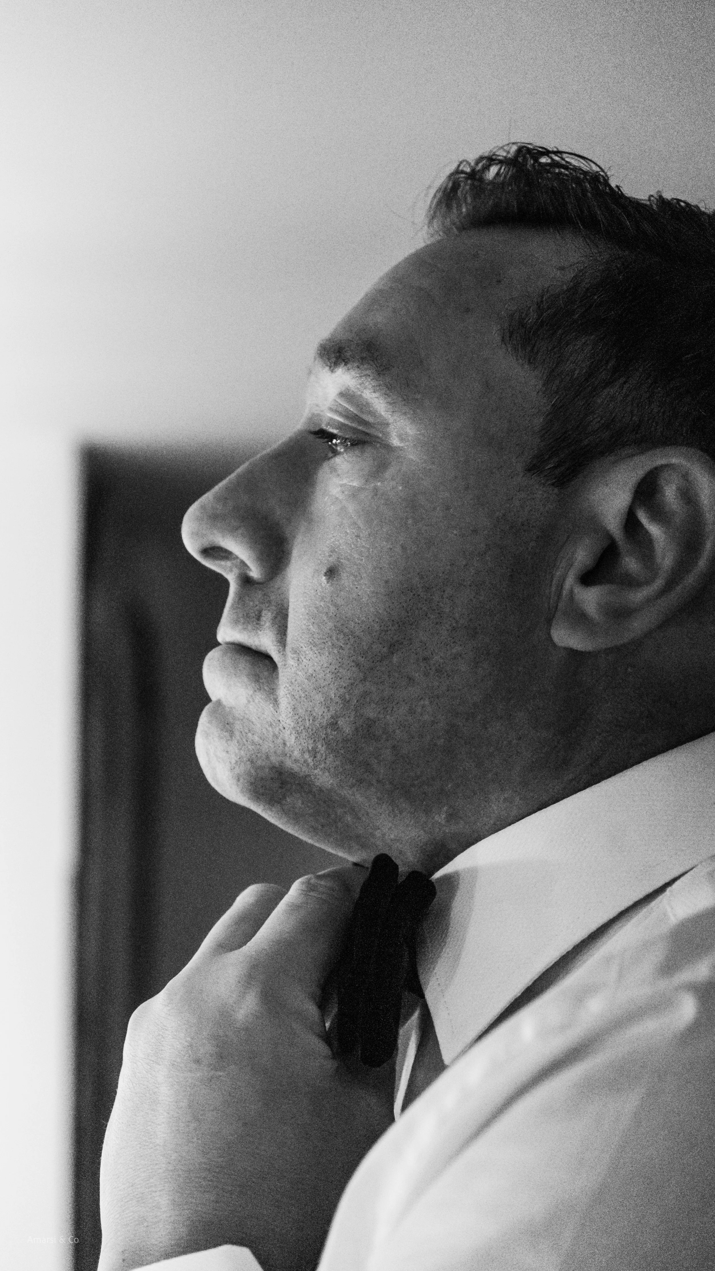 Close-up black-and-white photo of a man in formal attire, adjusting his bow tie.