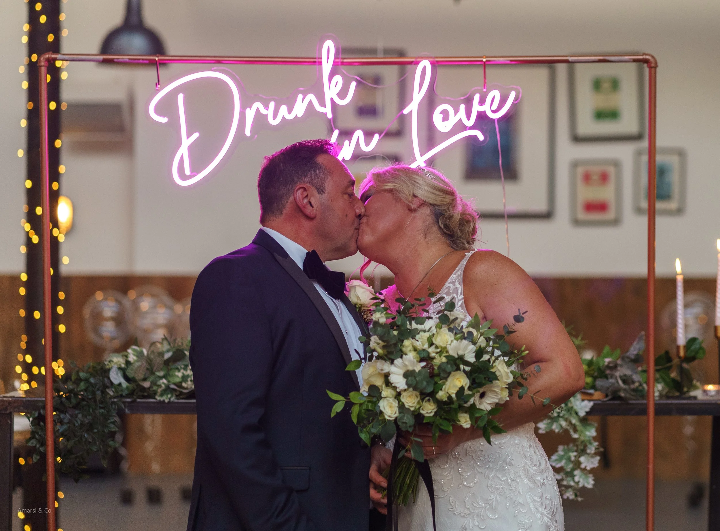 A couple in wedding attire sharing a kiss in front of a pink neon sign that reads 'Drunk in Love', with floral arrangements and candles in the background.