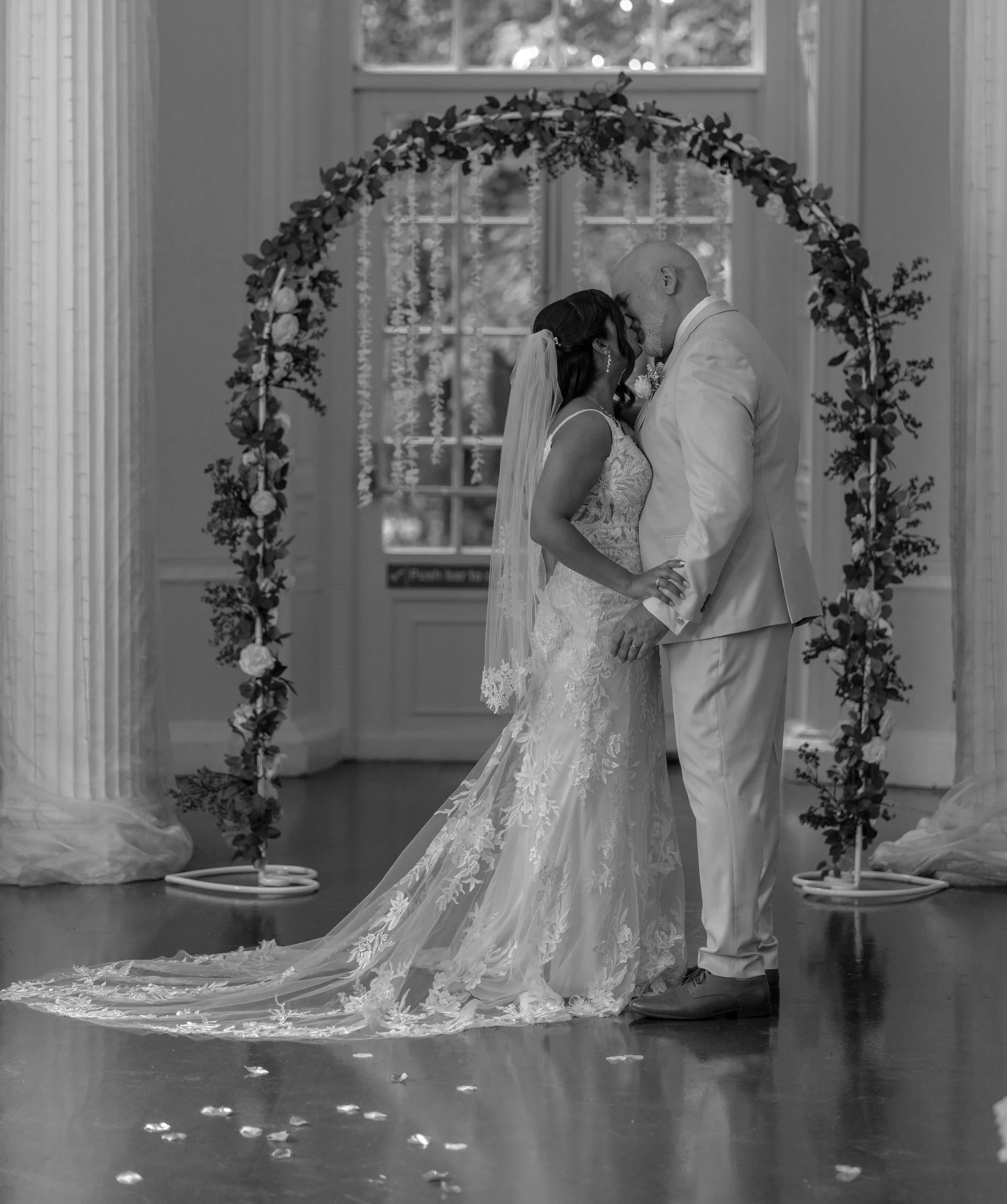A bride and groom sharing a kiss at their wedding ceremony, standing under a floral arch, with flower petals on the floor in front of them. Colwick hall wedding.