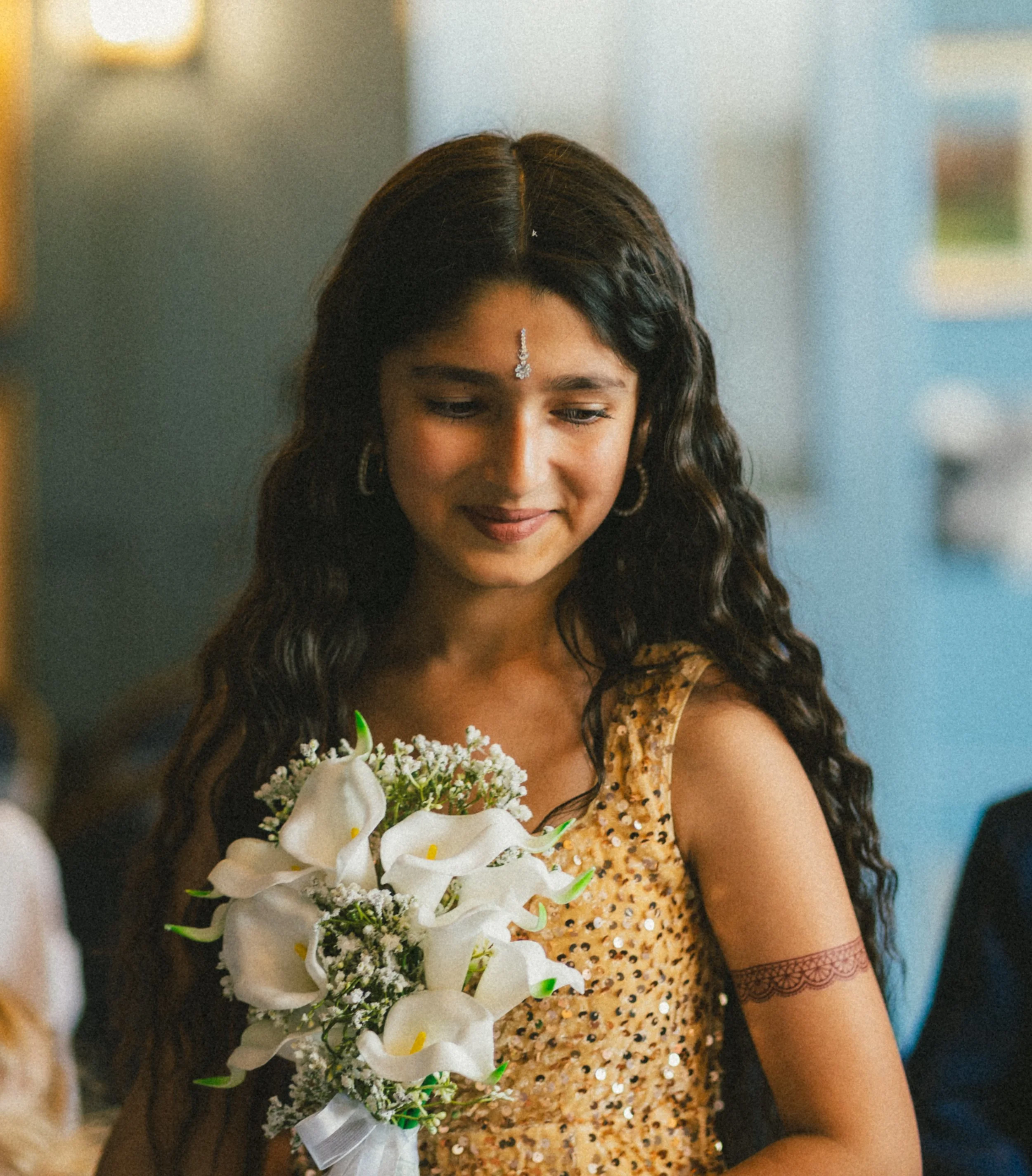 A woman with long dark curly hair, wearing traditional jewelry and a floral dress, holding a bouquet of white flowers.