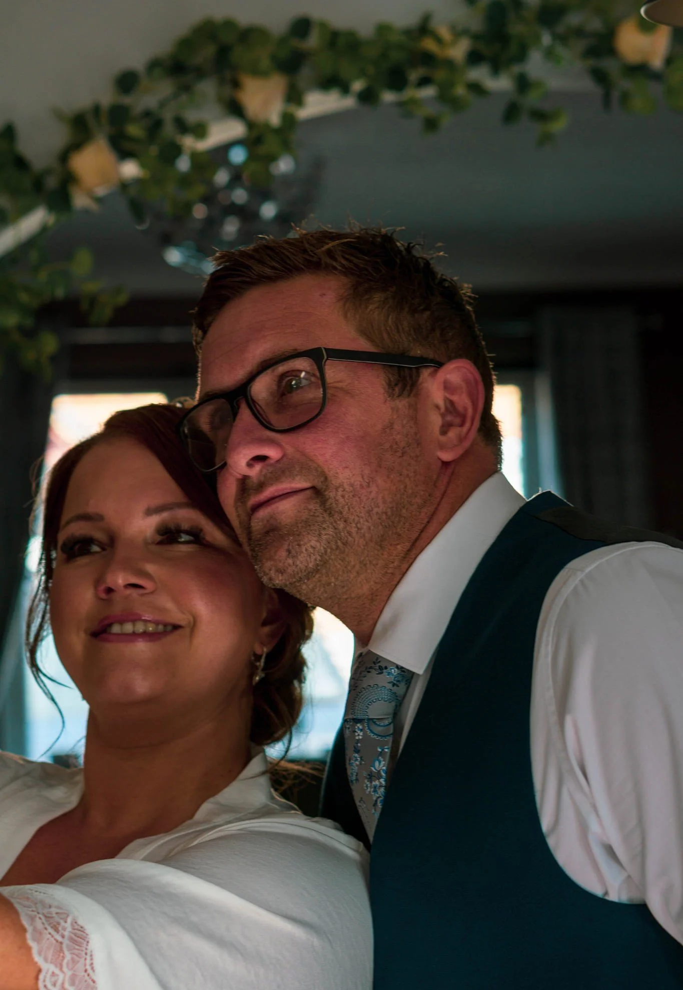 A man and a woman are hugging each other at a celebration. The woman has red hair and is smiling, while the man has short brown hair, glasses, and is wearing a suit with a patterned tie. They are indoors with decorative greenery hanging from the ceil