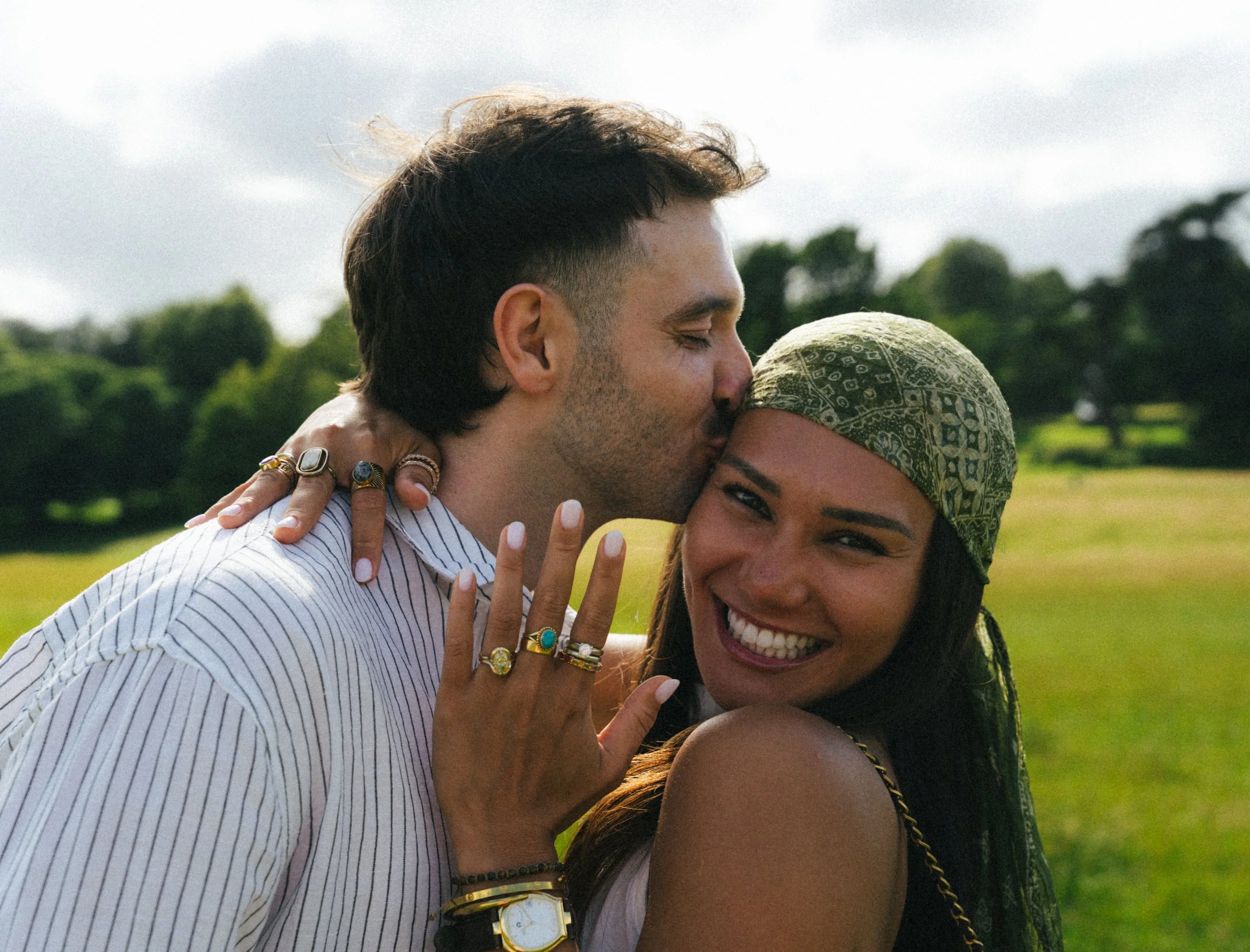 A man and a woman embrace outdoors, with the man holding the woman’s head close to his face, both smiling, with trees and a cloudy sky in the background.