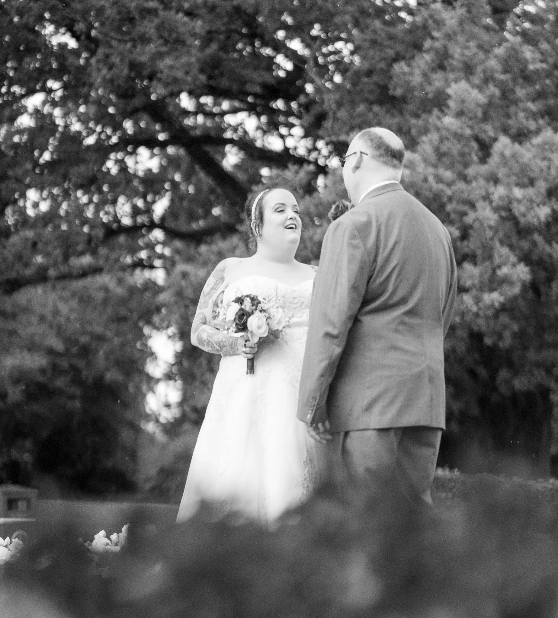 A black and white photograph of a bride and groom during their wedding ceremony outdoors. The bride is holding a bouquet of flowers, looking at the groom with a smile, and has tattoos on her arms. They are standing in front of a large tree with dense