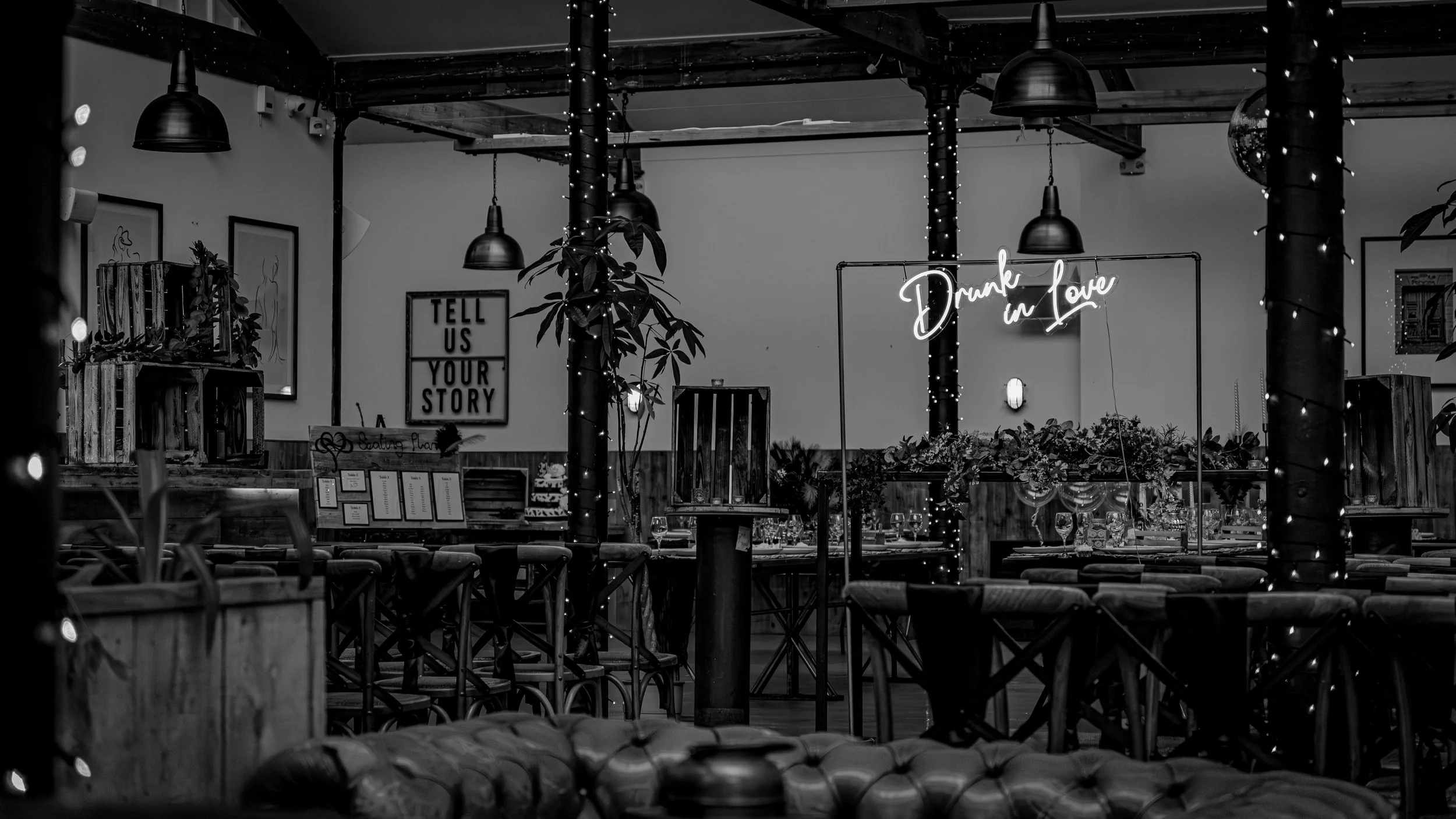 A dimly lit restaurant interior with wooden furniture, decorative plants, and string lights. A neon sign reads "Drunk in Love." There are framed wall art and a sign that says "Tell us your story." The atmosphere is cozy and romantic.